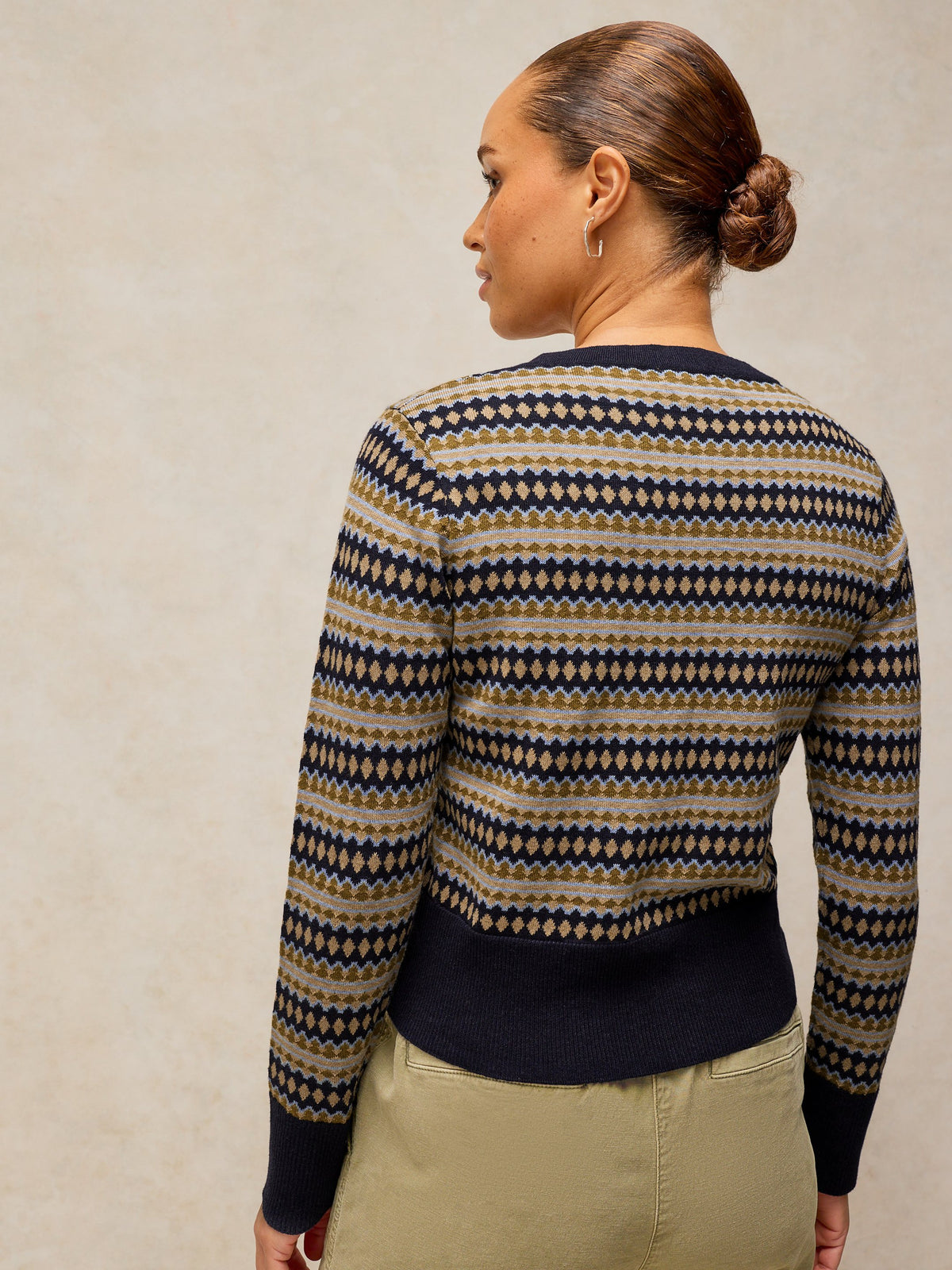 A woman shown from the back wearing a long-sleeve knit sweater with a horizontal geometric pattern in navy blue, beige, and brown, paired with khaki pants against a textured beige background.
