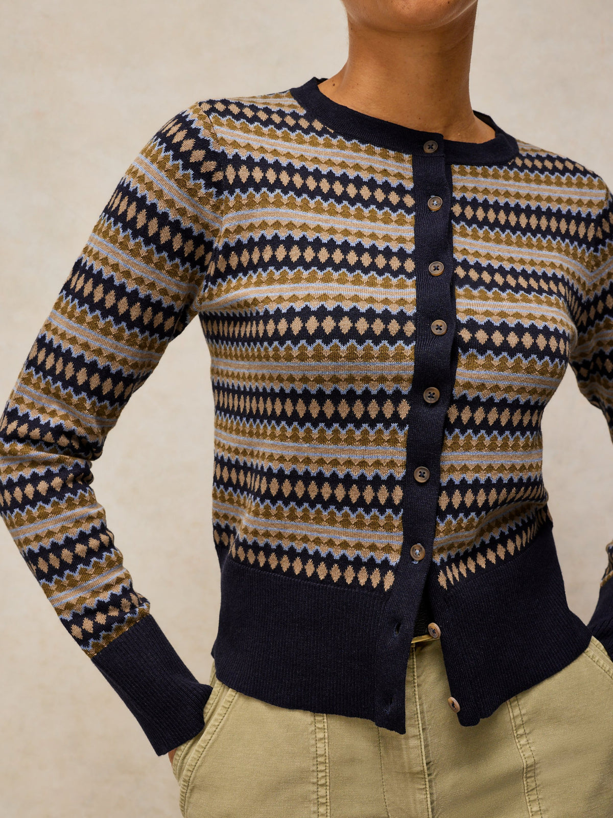 A close-up of a woman wearing a navy blue, beige, and brown geometric patterned cardigan sweater with a button-up front, paired with khaki pants.