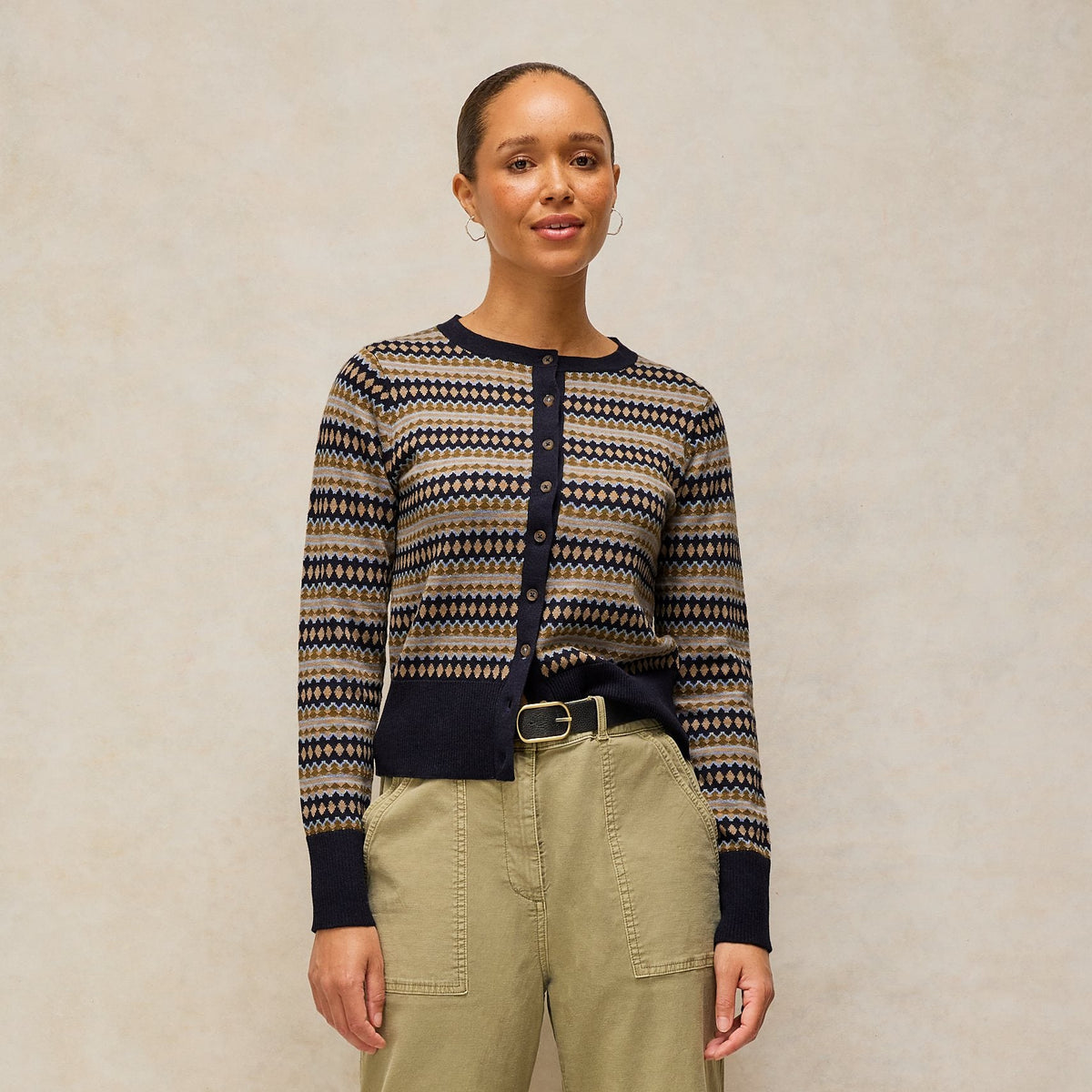 A woman modeling a navy and beige patterned knit cardigan sweater paired with olive green wide-leg trousers and a black belt, standing against a neutral textured beige backdrop.