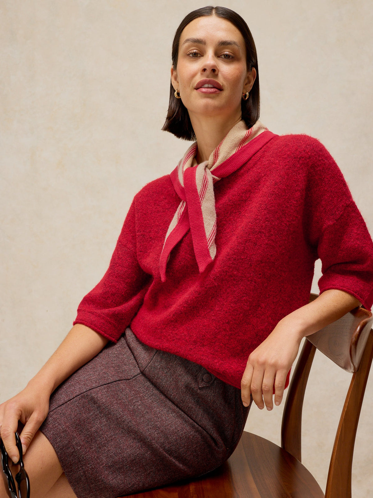 A woman seated on a wooden chair wearing a bright red knit sweater with a pink and white striped neck scarf, paired with a dark mauve skirt, against a neutral beige wall.