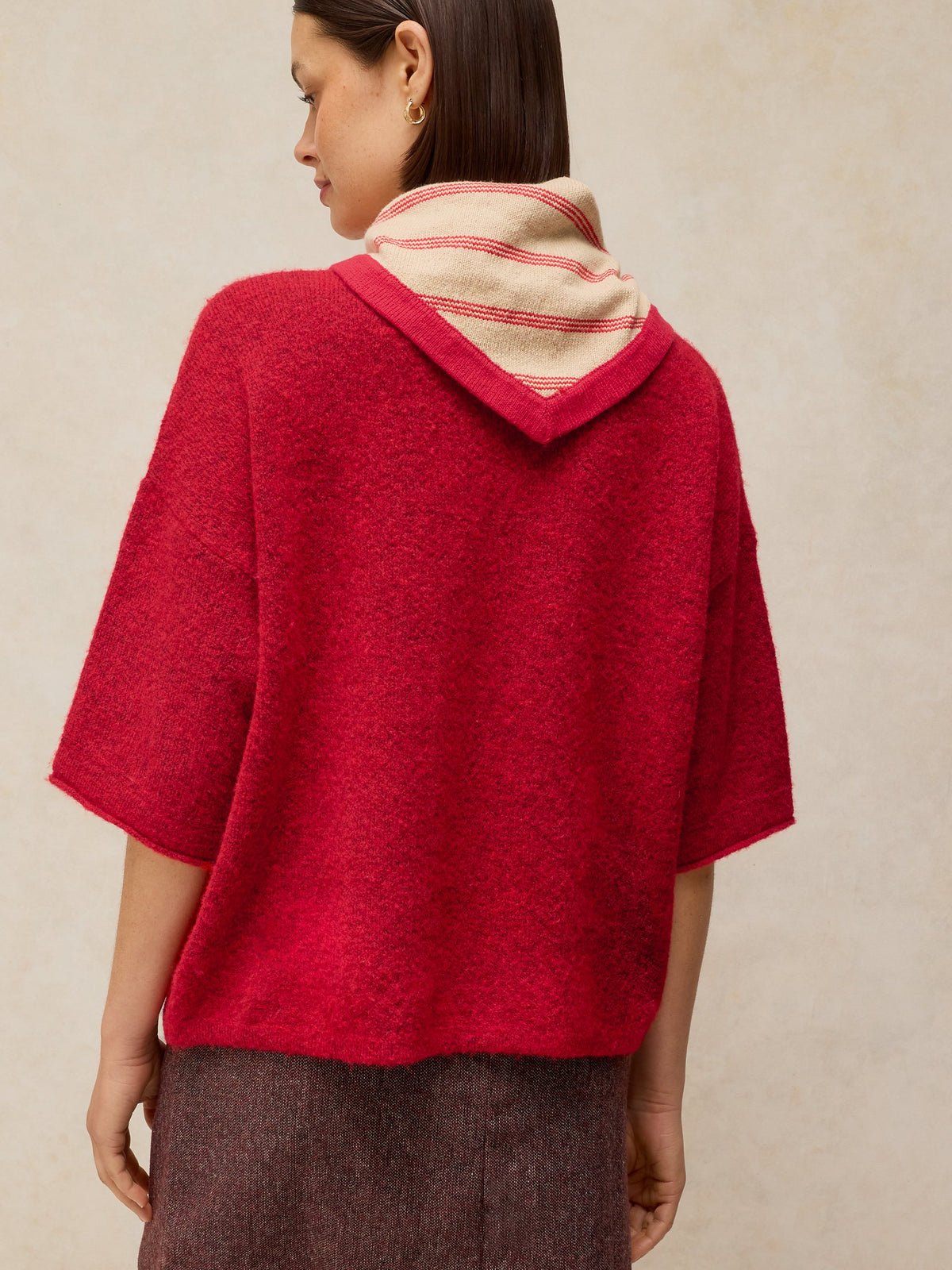 A woman shown from the back wearing a bright red fuzzy short-sleeve top with a beige and red striped scarf, paired with brown trousers, against a neutral beige background.