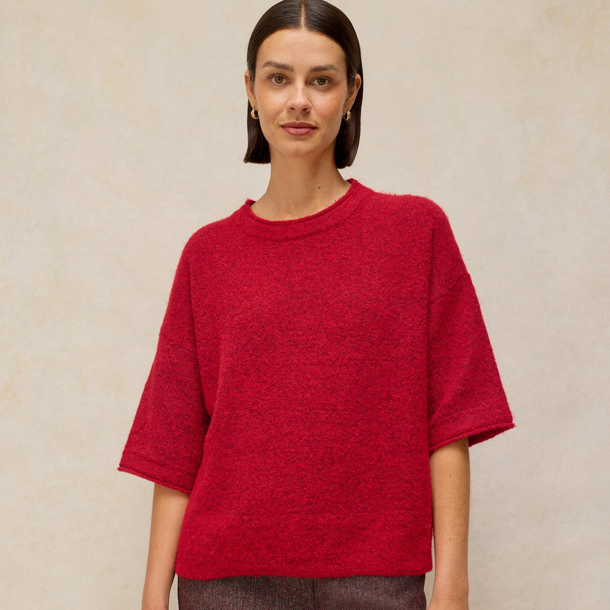 A woman modeling an oversized short-sleeve knit top in heathered red, paired with a textured dark brown mini skirt, against a neutral beige background.