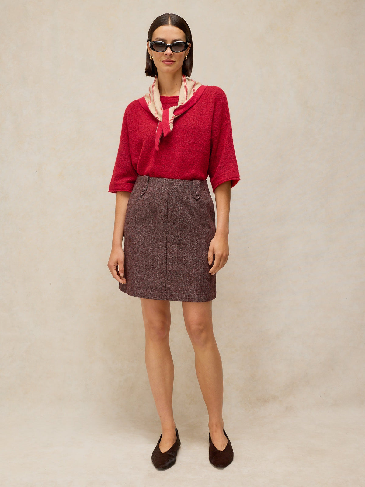 A woman stands against a plain beige studio backdrop wearing a bright red short-sleeve knit top with a light pink and red scarf tied around the collar, a brown textured mini skirt, and dark brown pointed flats.