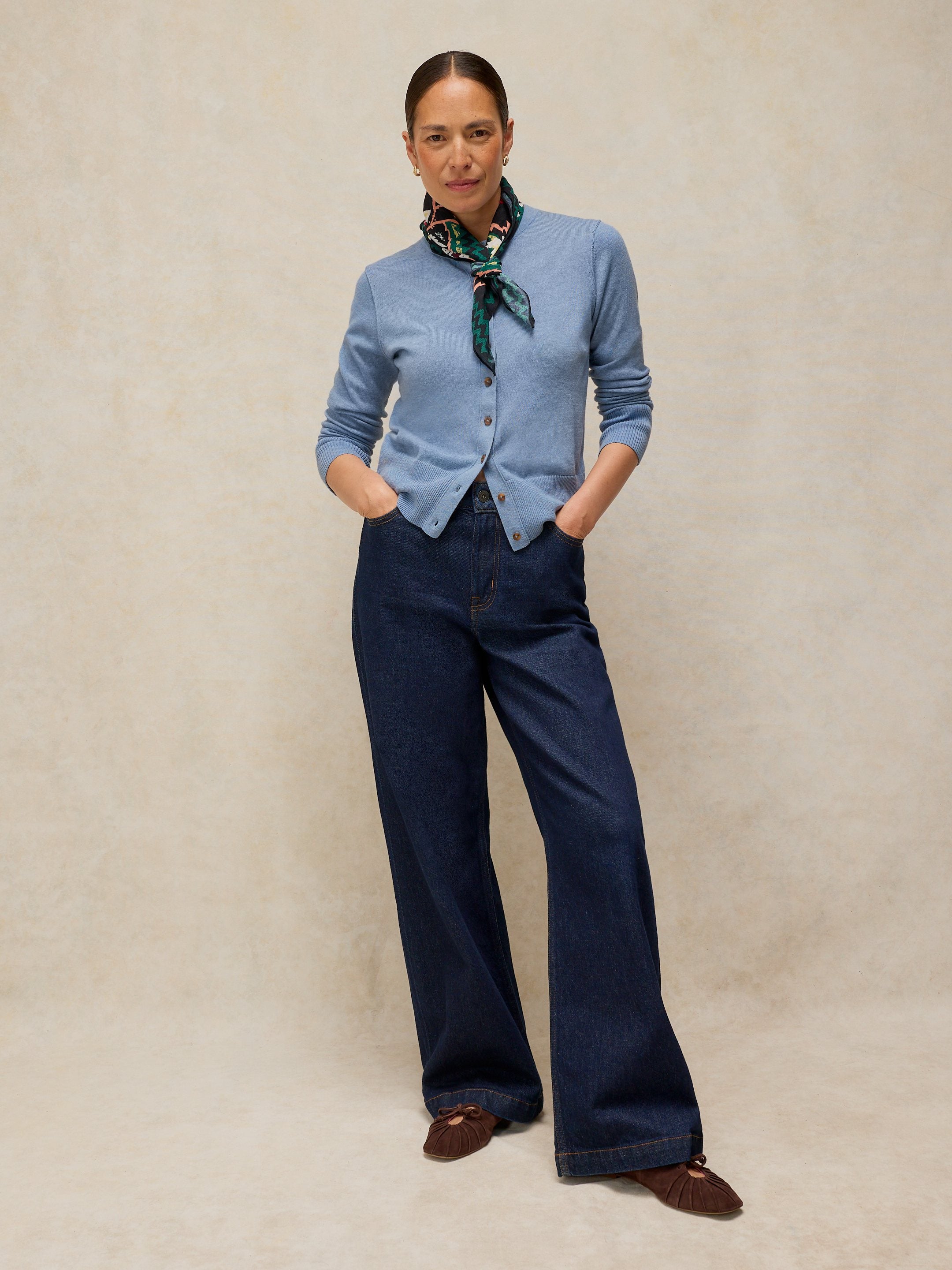 Woman wearing a light blue cardigan, dark blue pants, and brown shoes against a beige background