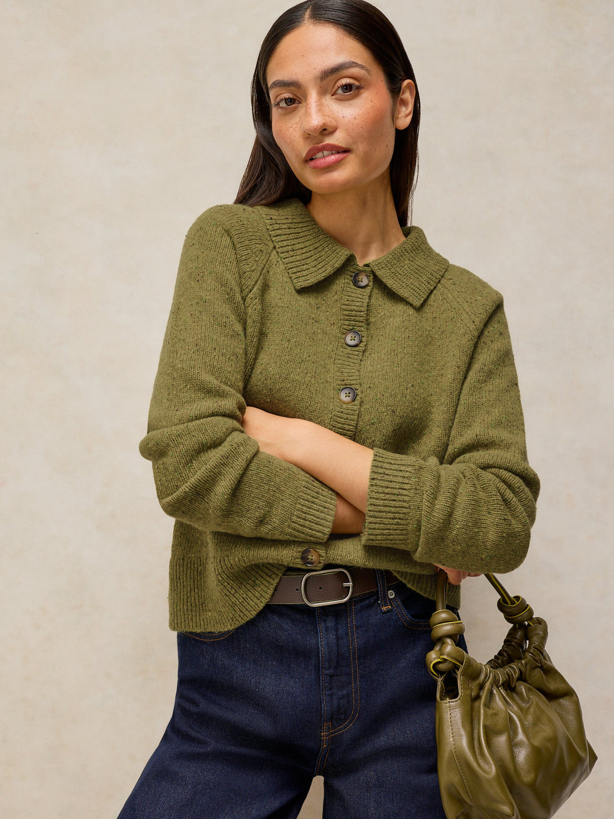 A woman wearing an olive green knit button-front cardigan with a collar, paired with dark blue jeans and holding an olive green handbag, set against a neutral textured beige background.
