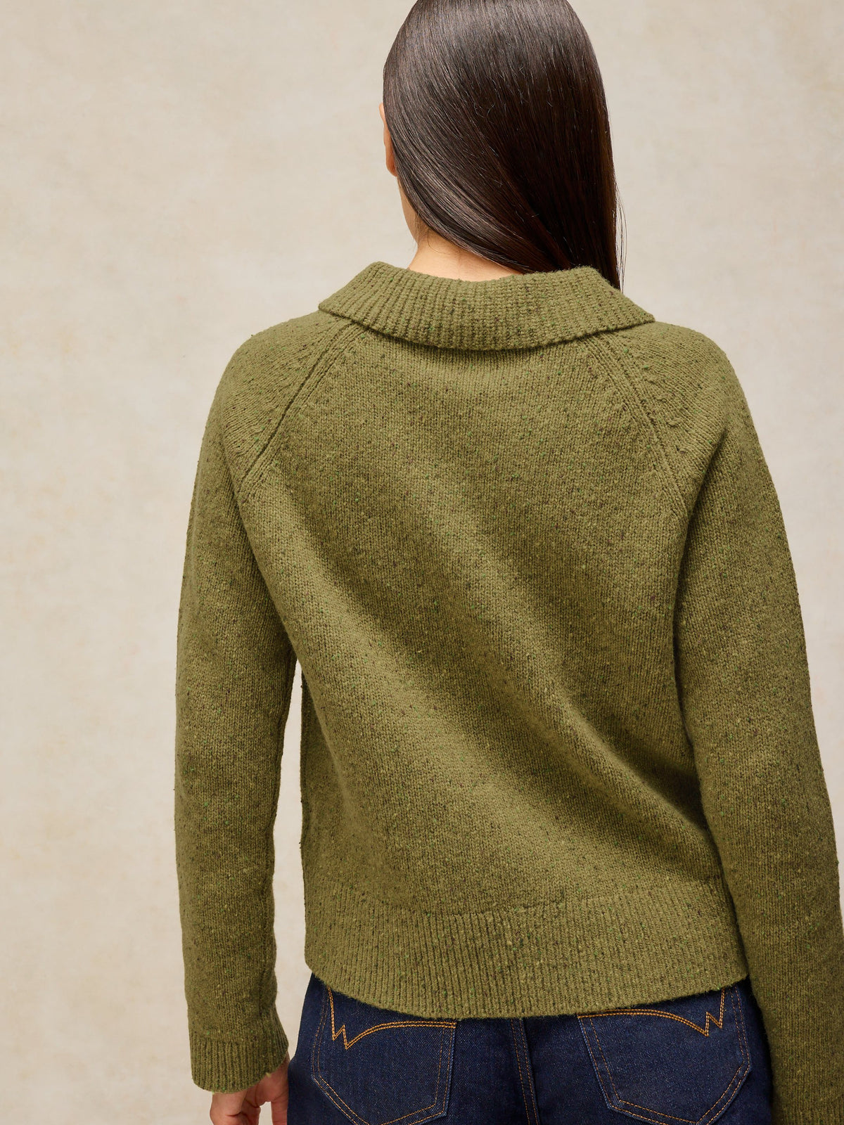 A woman viewed from the back wearing an olive green knit sweater with raglan sleeves and a ribbed mock neck, paired with dark blue jeans against a textured beige background.