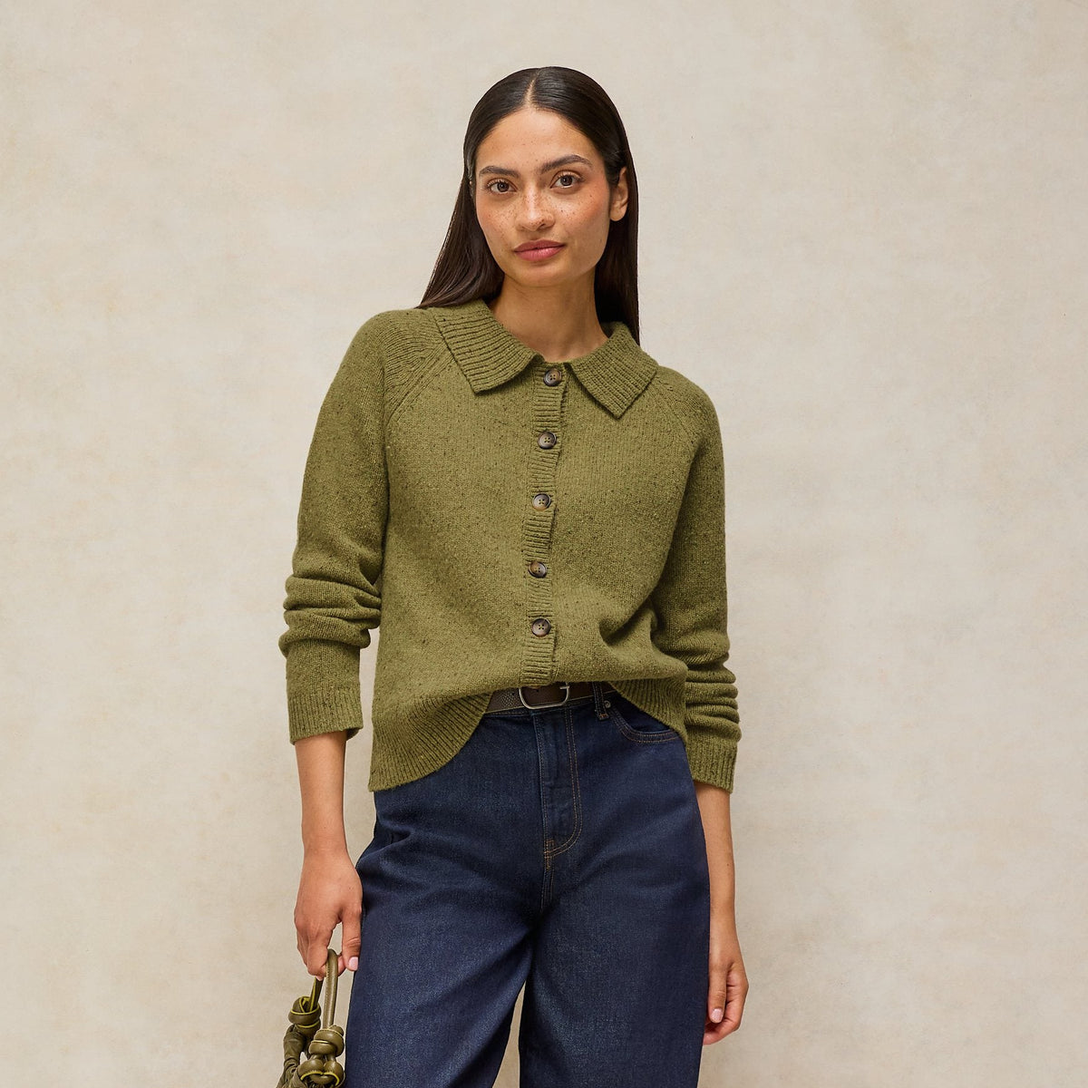 A woman stands against a warm beige textured backdrop wearing an olive green knit button-front top, dark blue jeans, burgundy flats, and holding a small olive green handbag.