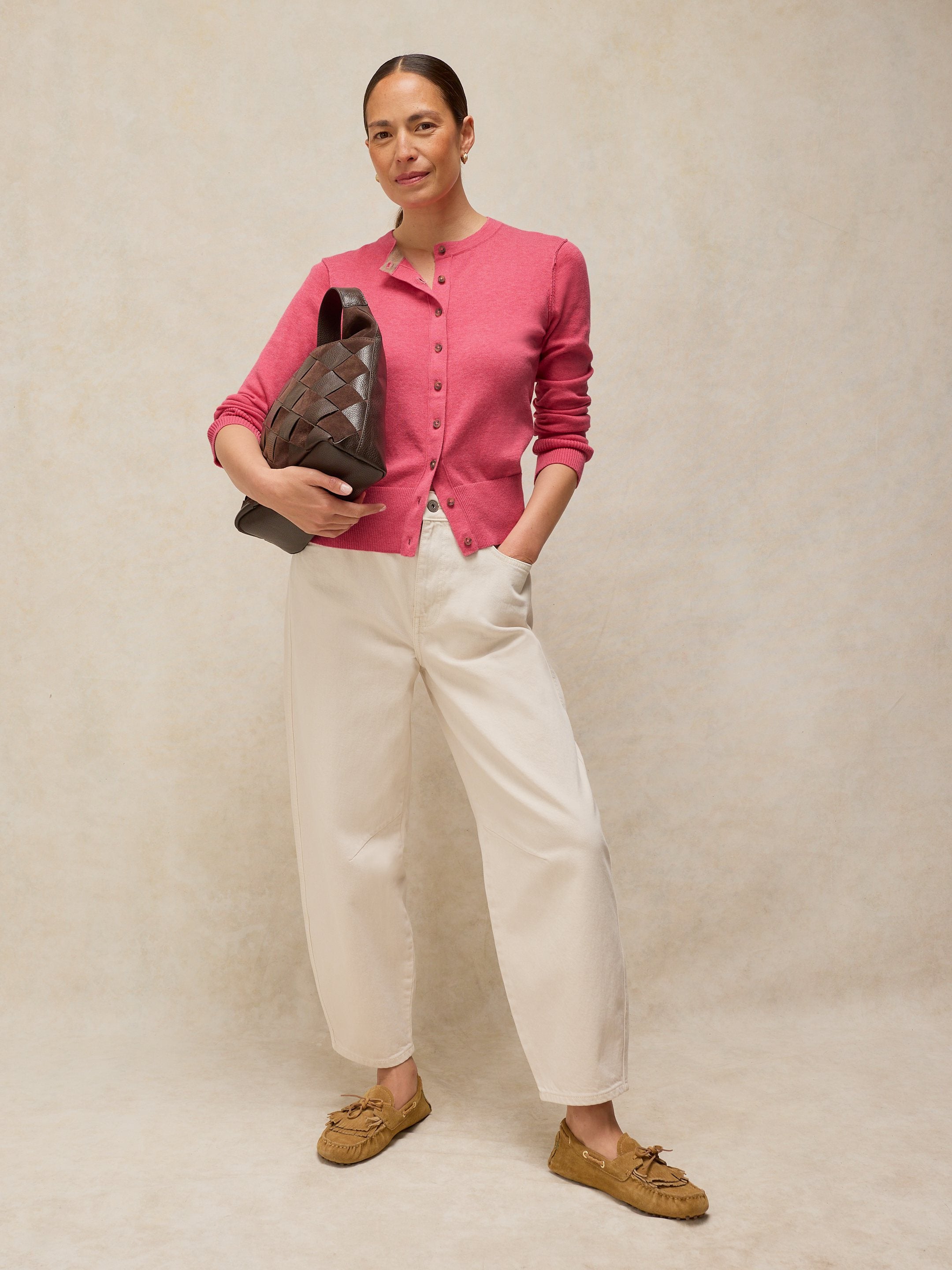 Woman wearing a pink shirt, white pants, and brown shoes holding a brown bag against a beige background