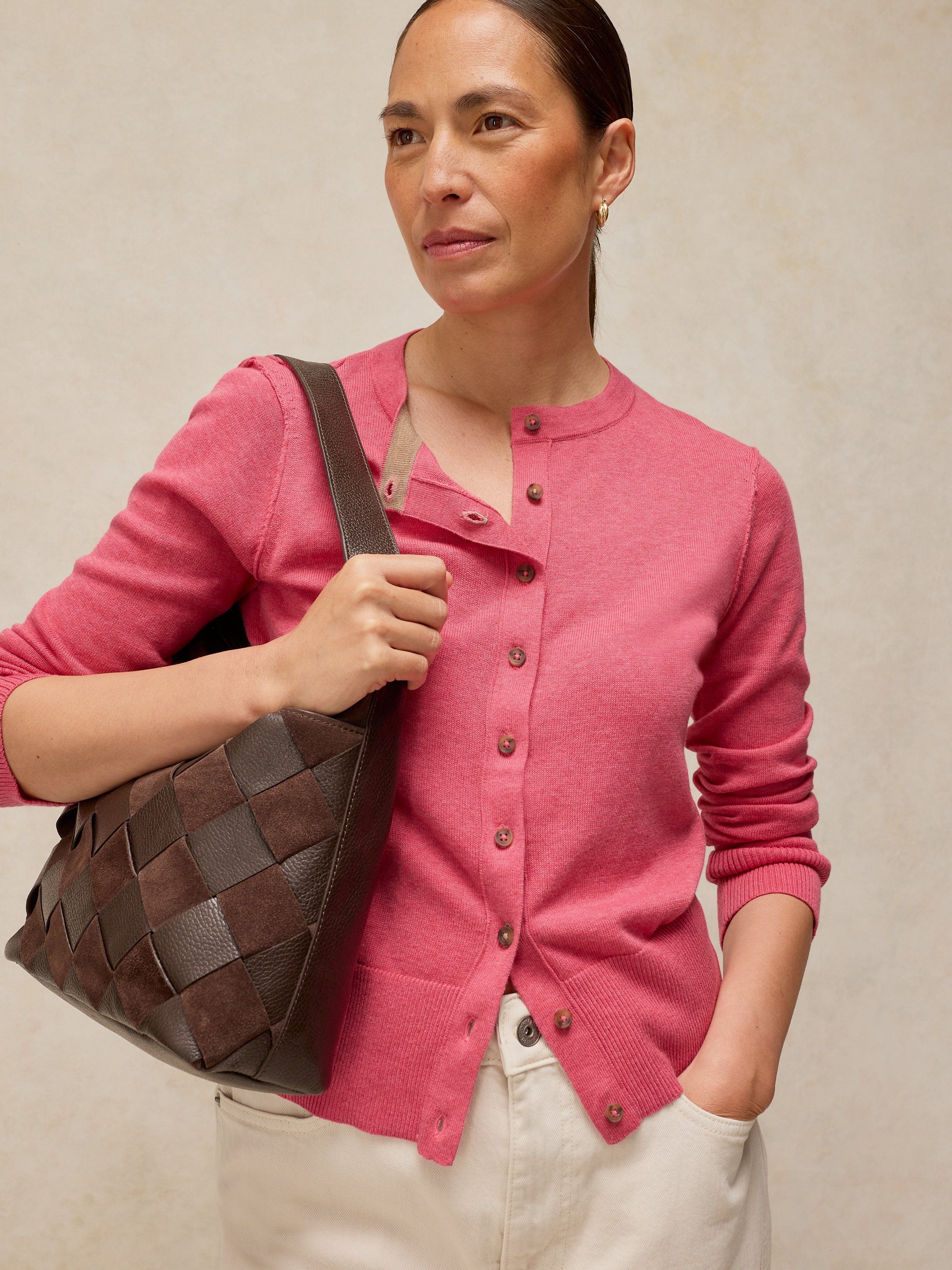 Woman wearing a pink cardigan holding a brown woven handbag against a beige background