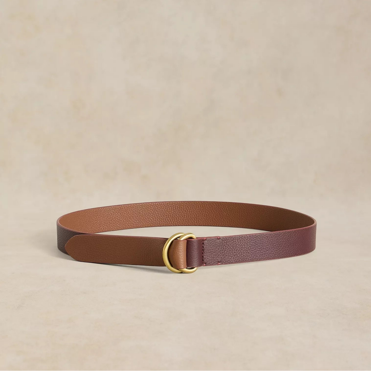 A slim leather belt in a two-tone brown design with a gold-tone metal ring buckle, displayed on a textured beige background.