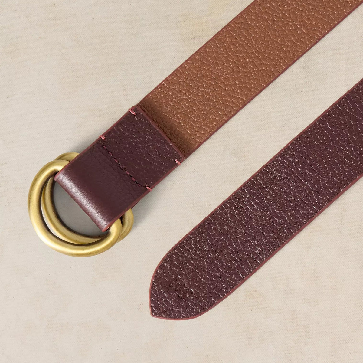 A close-up of a brown pebbled leather belt with a wide burgundy double-ring metal buckle and a matching burgundy leather keeper, laid diagonally on a light beige textured background.
