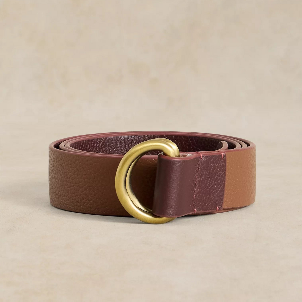 A brown leather belt with a color-blocked design featuring a darker brown outer strap, a reddish-brown inner strap, and a large polished gold-tone O-ring buckle, displayed on a neutral beige background.