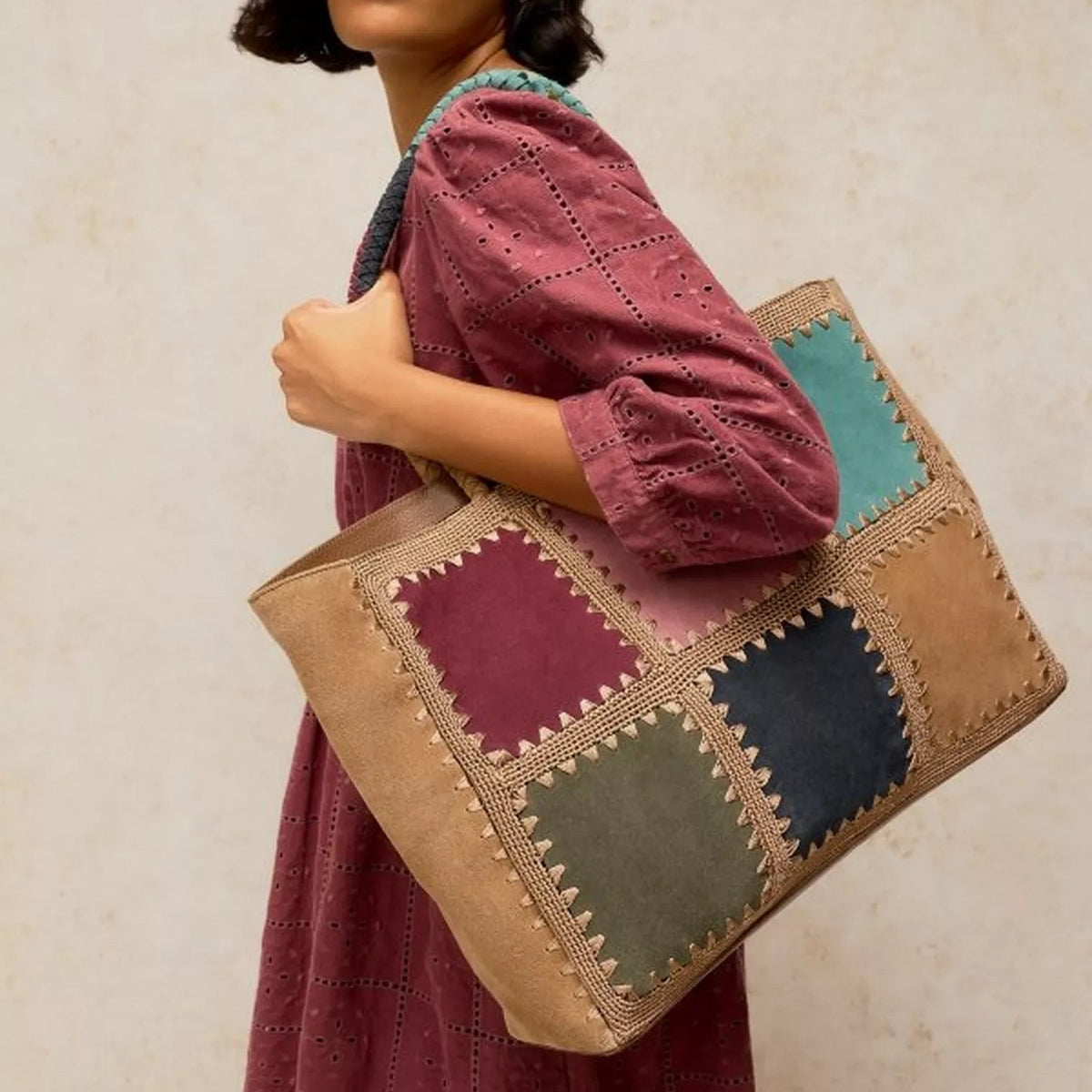 A woman wearing a dusty rose eyelet dress carries a large patchwork tote bag. The bag features square panels in tan, burgundy, teal, olive green, and navy blue with decorative stitching.