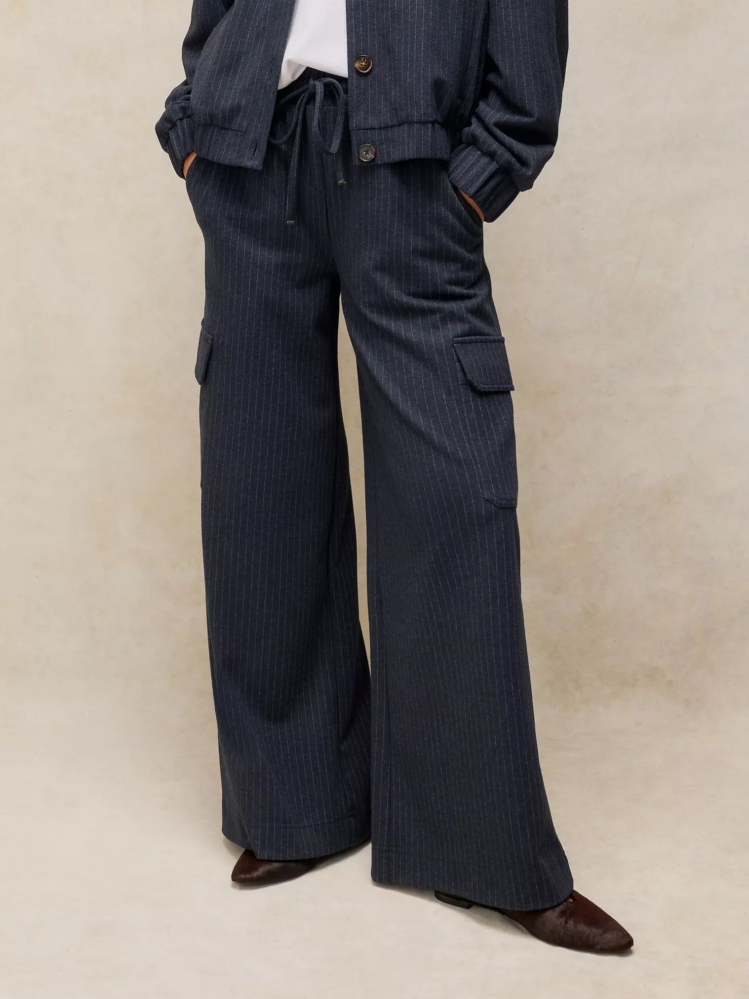 A person wearing dark navy pinstriped wide-leg trousers with cargo pockets, paired with a matching jacket and brown pointed-toe shoes, against a neutral beige studio backdrop.