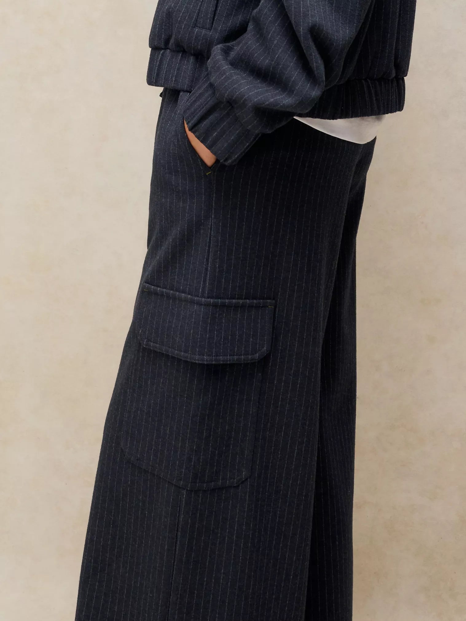 Close-up of a person wearing dark navy pinstriped wide-leg trousers with a large front flap pocket, paired with a matching dark ribbed top against a warm beige textured background.
