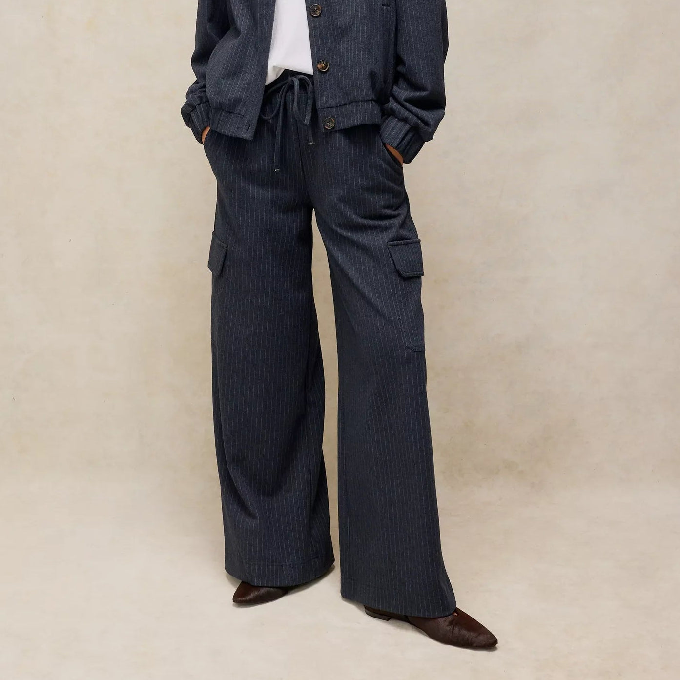 A woman stands against a textured beige studio backdrop wearing a dark navy pinstriped jacket and matching wide-leg cargo pants over a white top and brown shoes.