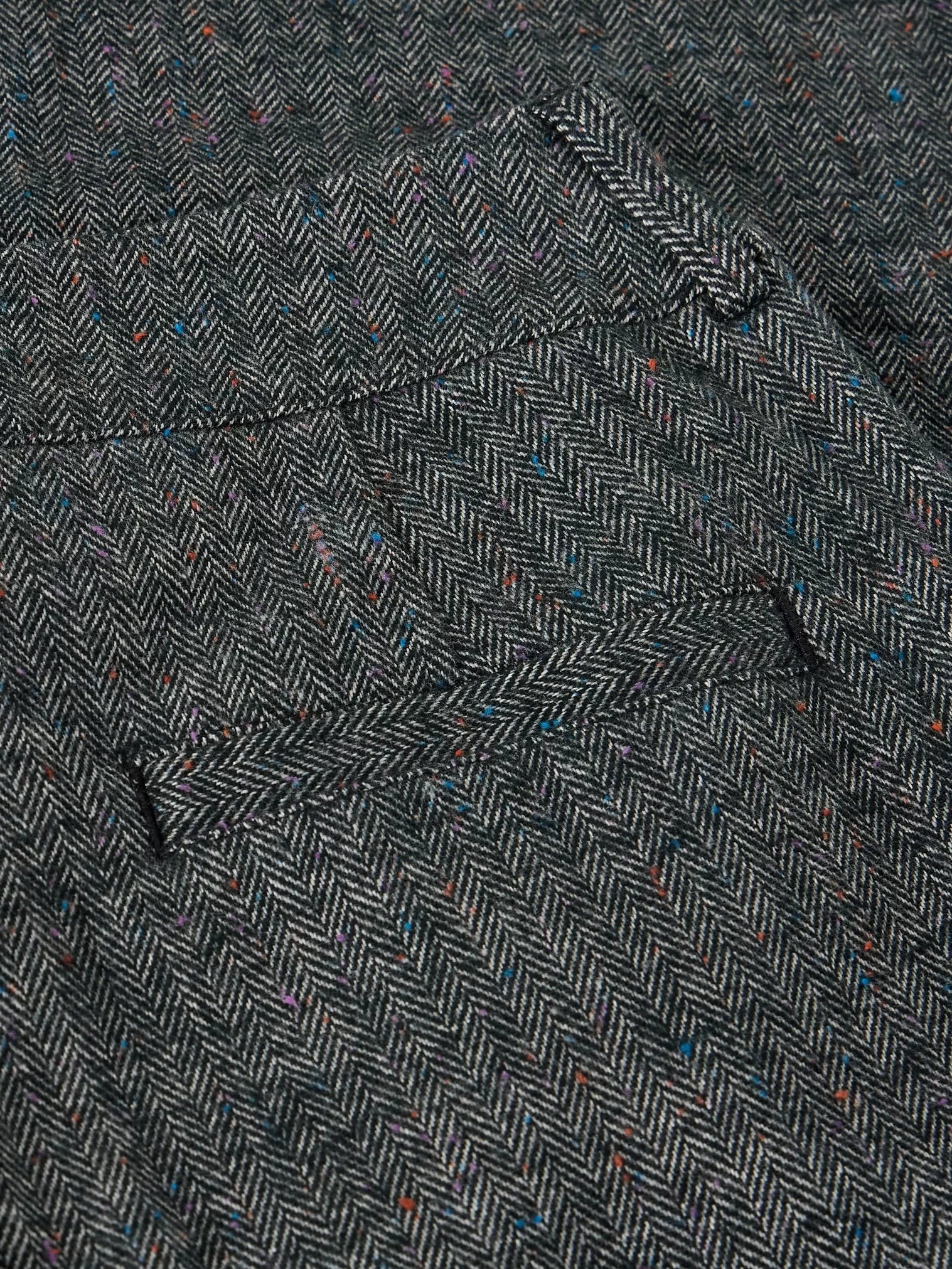 Close-up of a dark gray and black herringbone tweed fabric with subtle multicolored flecks, showing a pocket and buttonhole detail.