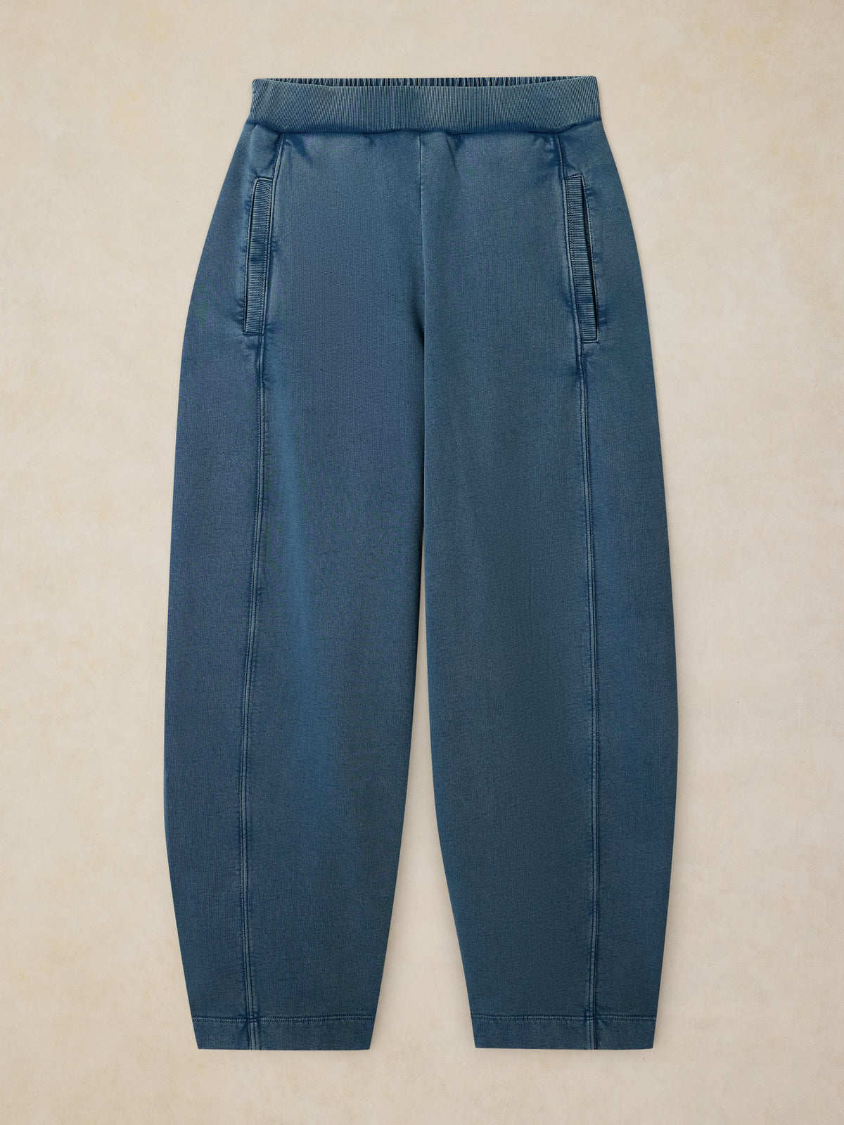 A pair of relaxed-fit denim trousers in a muted slate blue color, laid flat against a textured beige background.