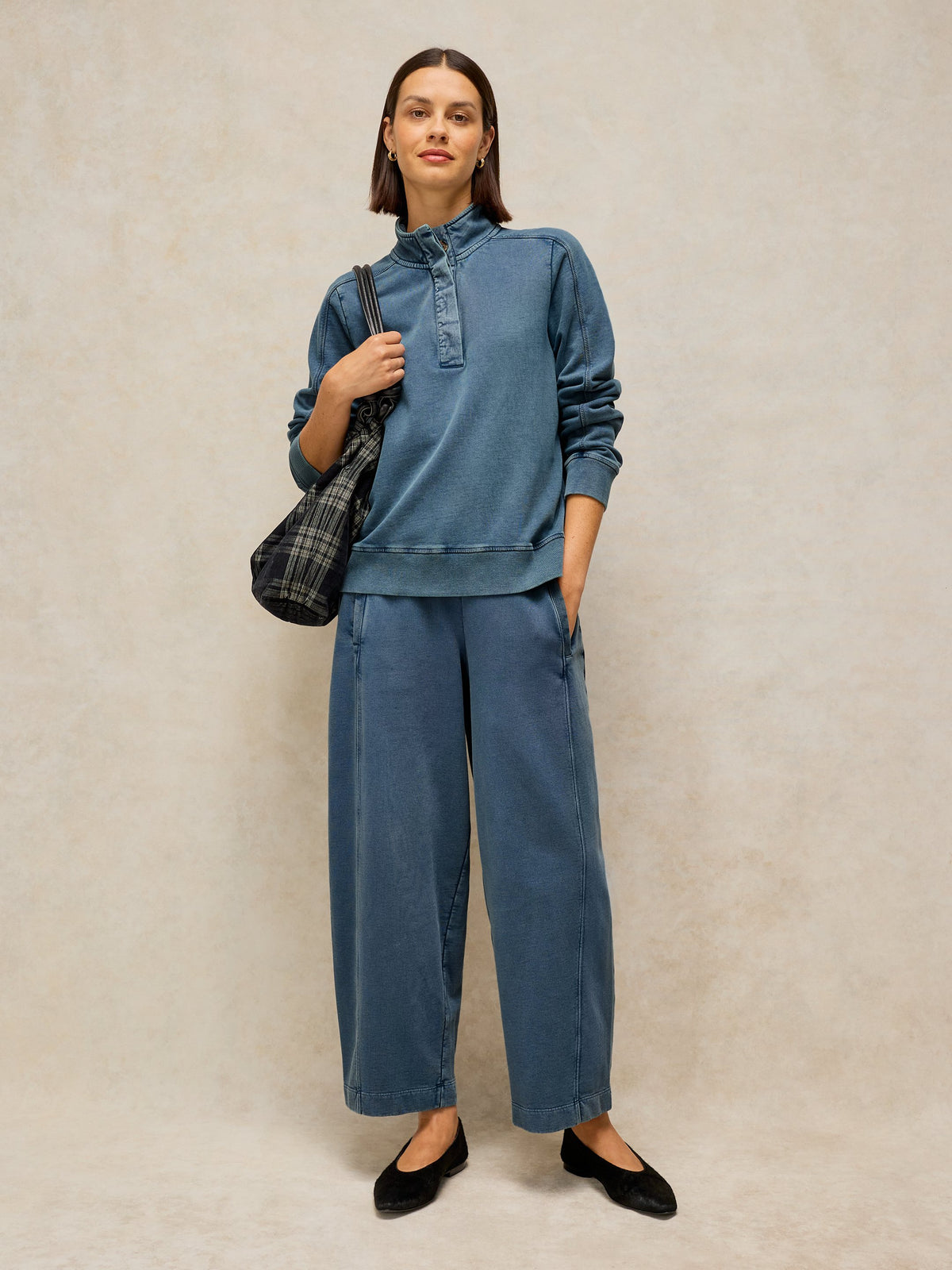 A woman modeling a matching blue denim lounge set consisting of a long-sleeve mock-neck top and wide-leg pants, styled with a plaid shoulder bag and black flats against a textured beige studio backdrop.