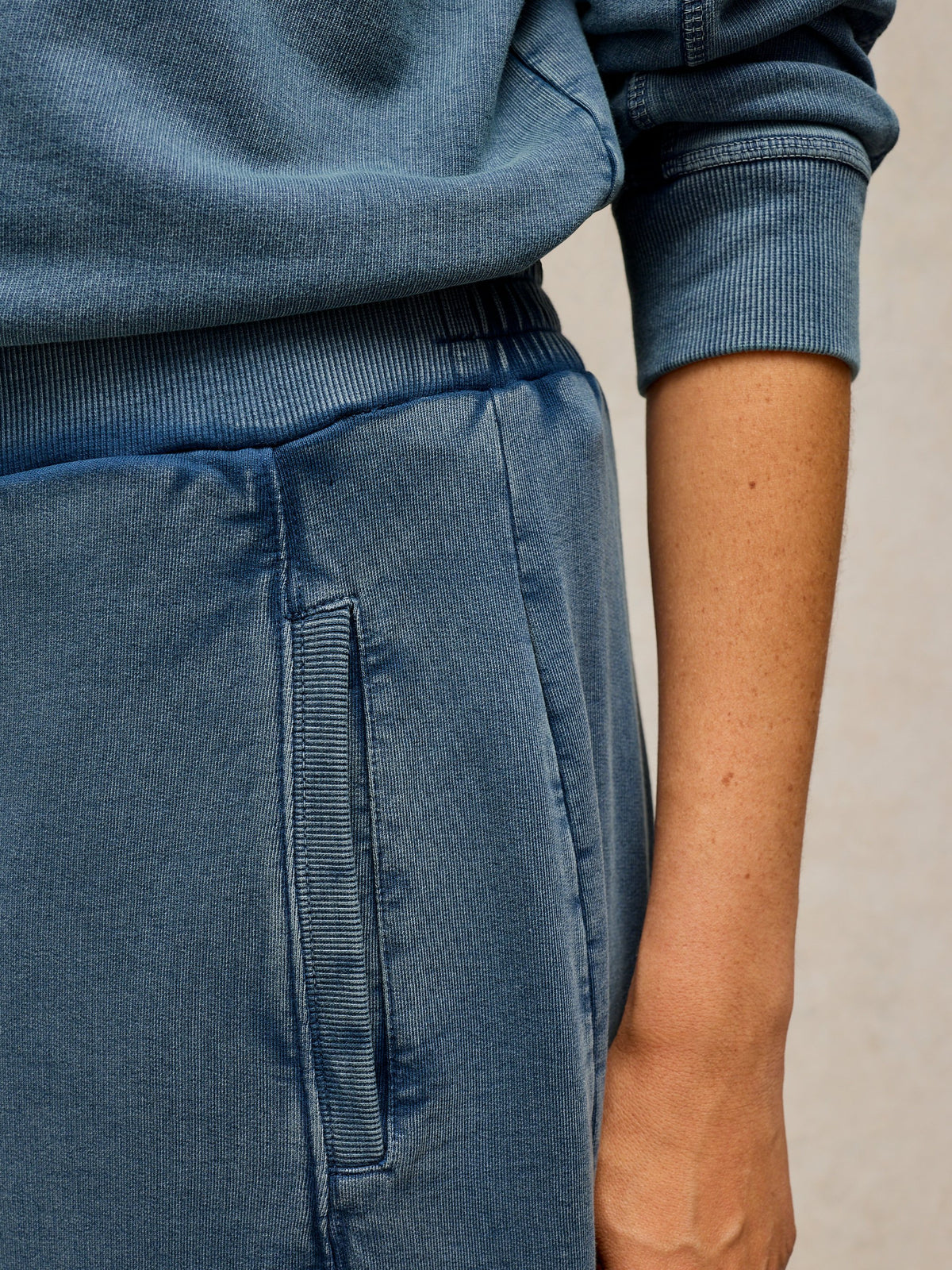Close-up of a person wearing a blue denim lounge set, featuring a ribbed waistband and a matching vertical welt pocket on the pants.