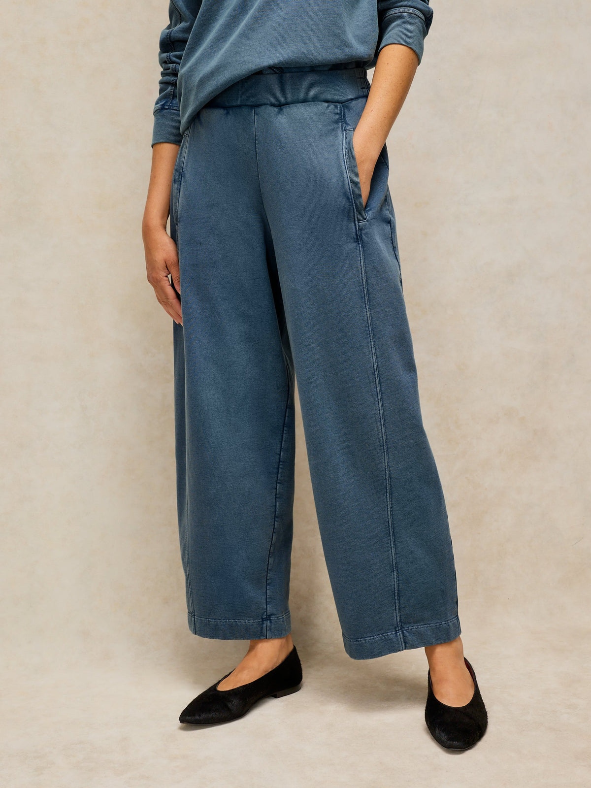 A person wearing wide-leg cropped pants in a muted blue denim-like fabric, paired with a matching blue top and black flats, standing against a warm beige textured backdrop.