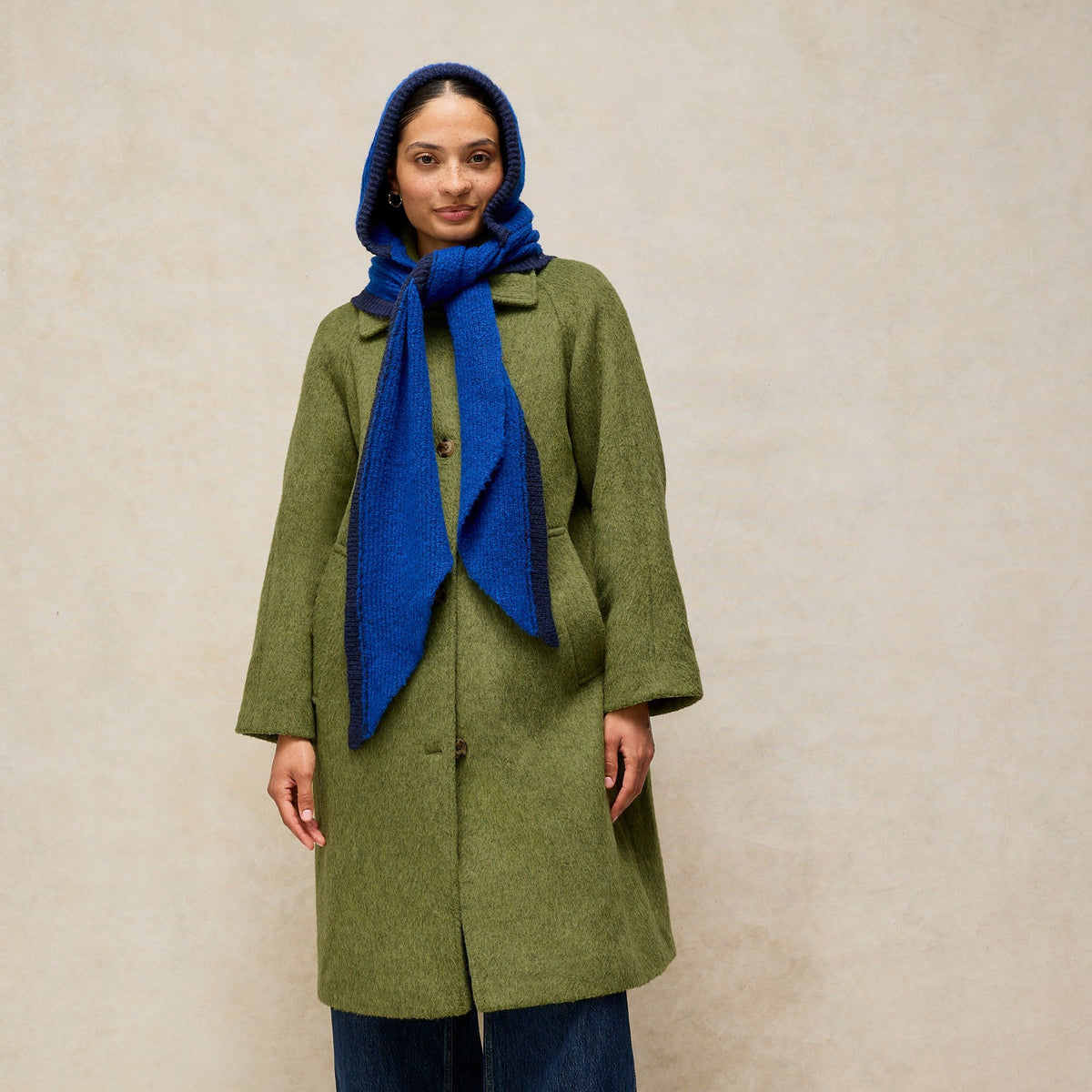 A model stands against a textured beige studio backdrop wearing a mid-length olive green coat, a bright blue knit headscarf and matching scarf, cuffed denim jeans, and brown leather loafers.