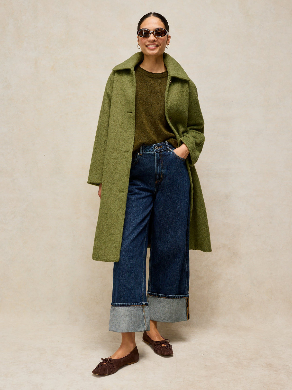 A woman modeling a long olive green coat over a dark olive top, blue wide-leg jeans with cuffed hems, and brown loafers, standing against a textured beige studio backdrop.