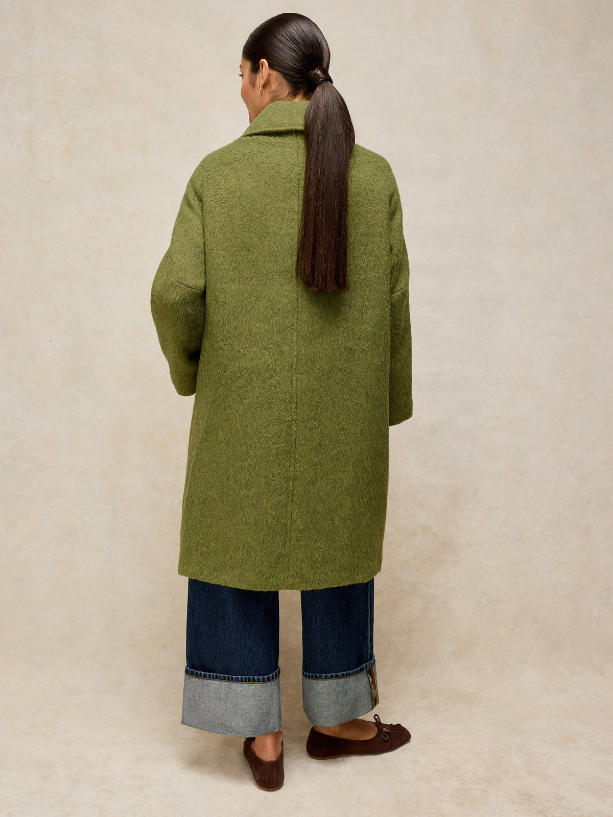 A woman viewed from behind wearing a long olive green wool-blend coat over dark blue cuffed jeans and brown flat shoes, standing against a textured beige studio backdrop.