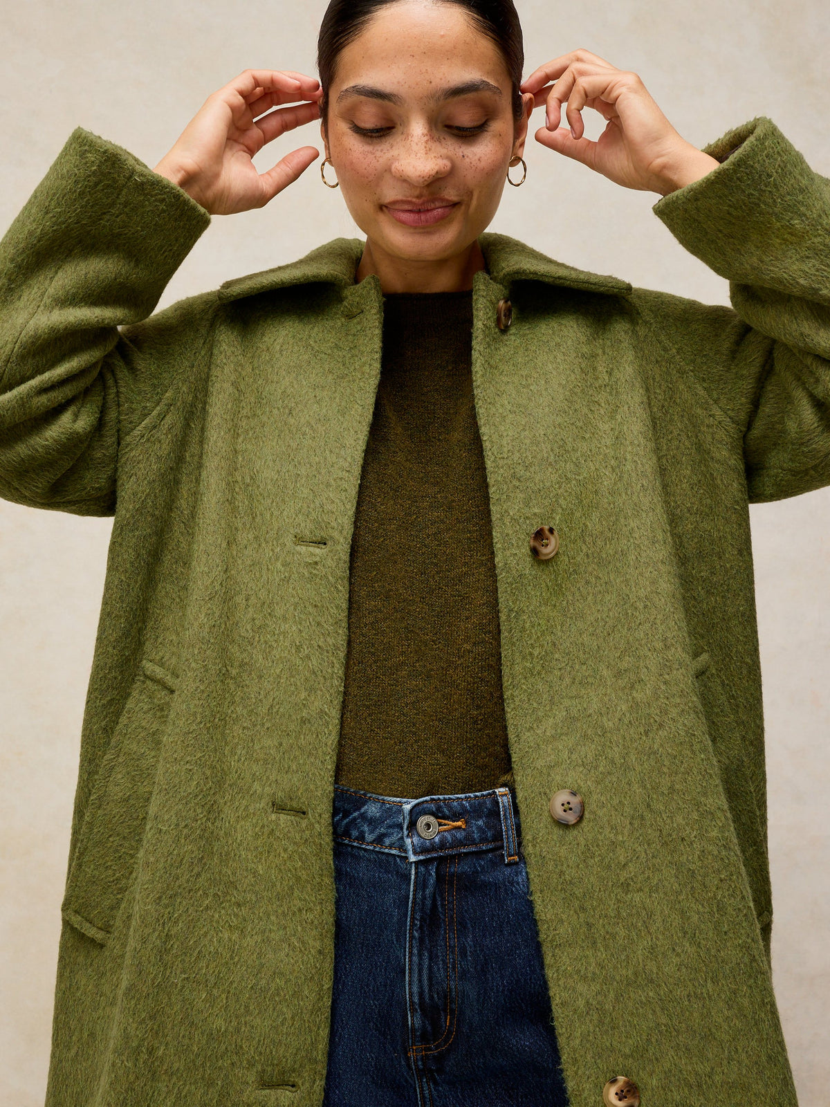A woman wearing a textured olive green wool-blend coat over a dark olive sweater and blue jeans, posing against a neutral beige background.