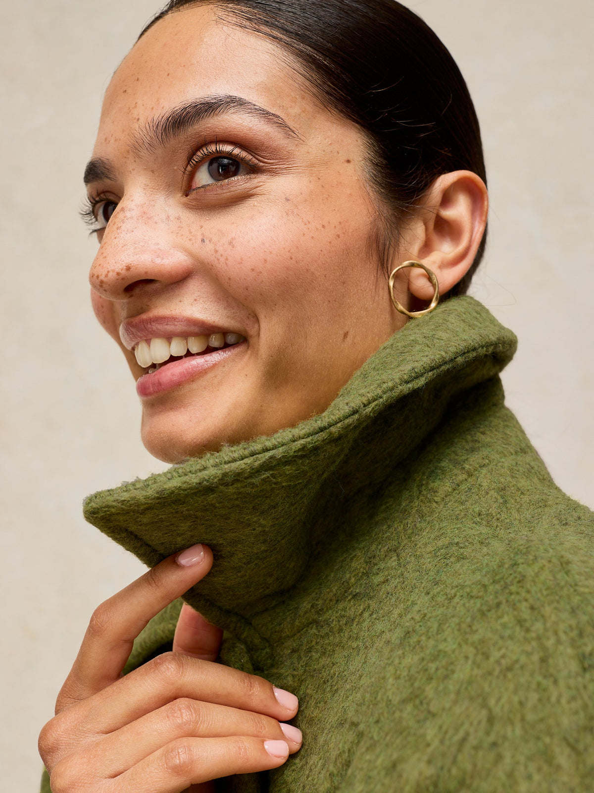 A close-up of a smiling woman wearing a textured olive green high-neck sweater and gold hoop earrings, set against a neutral beige background.