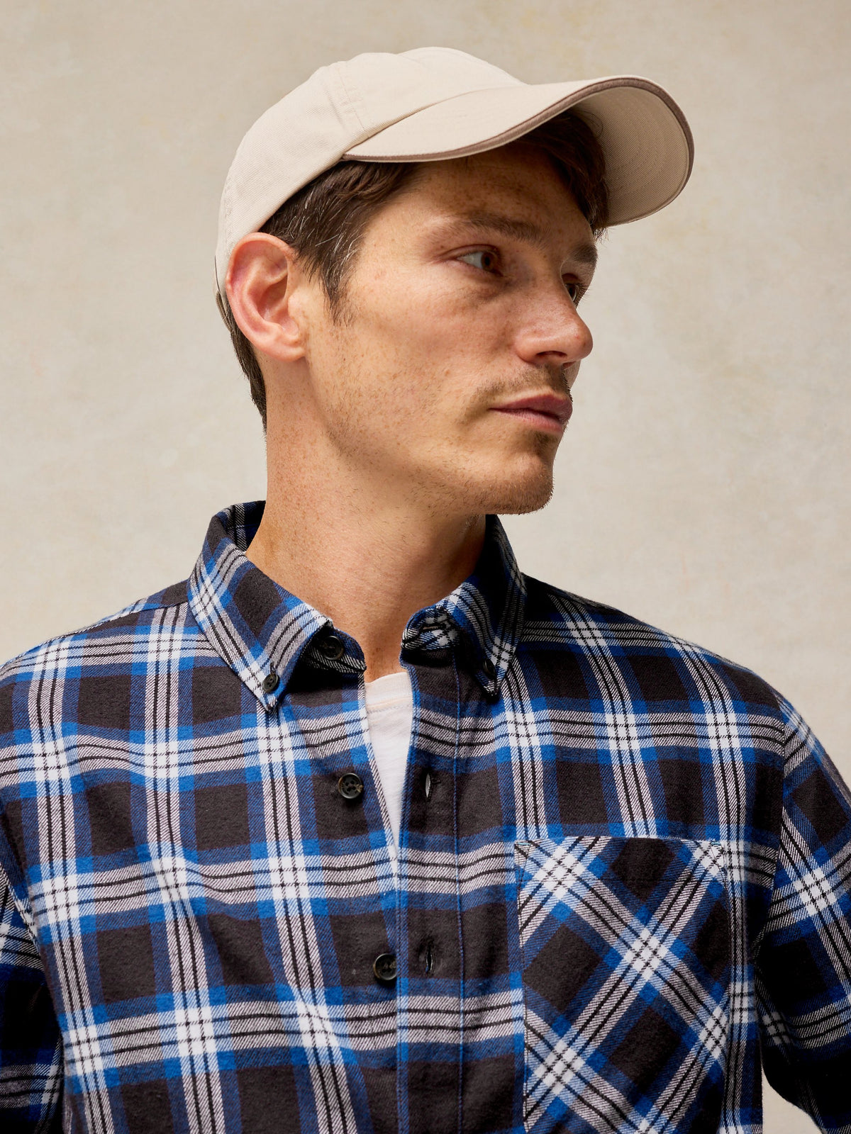 A man wearing a beige baseball cap and a blue, black, and white plaid button-down shirt, photographed against a neutral textured beige background.
