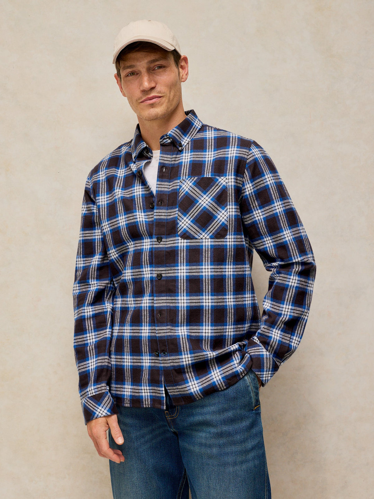A man wearing a navy blue and black plaid button-down shirt with white and light blue stripes, paired with blue jeans and a beige baseball cap, standing against a textured beige wall.