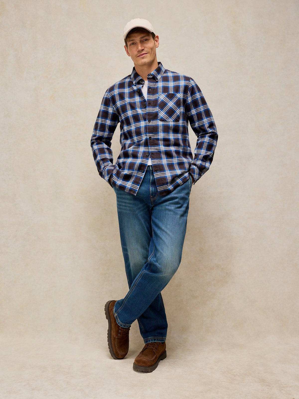 A man wearing a blue and black plaid button-down shirt, blue jeans, brown boots, and a beige baseball cap, standing against a textured beige studio backdrop.