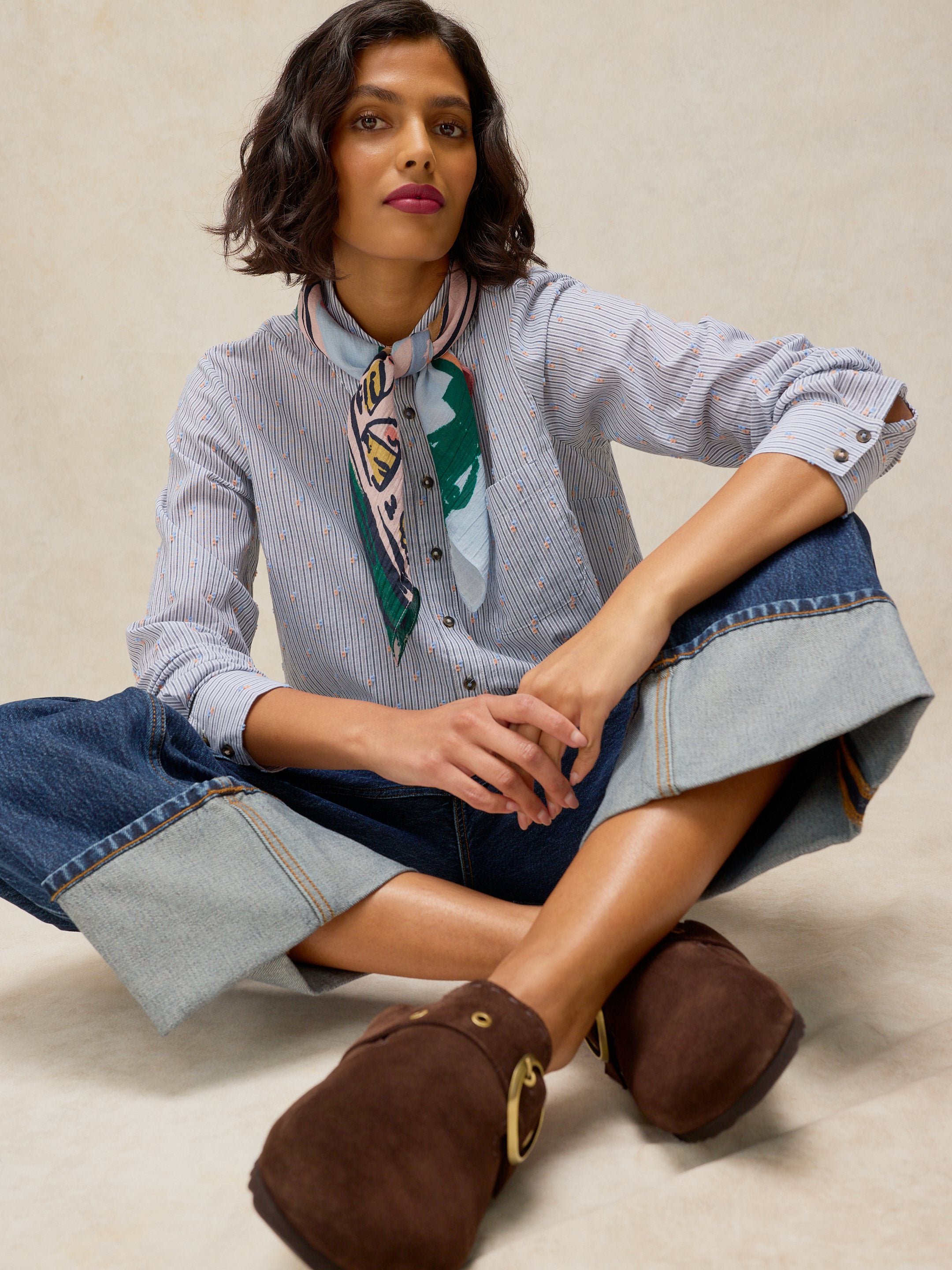 A woman sits cross-legged wearing a light blue and white striped button-up shirt with a green and white patterned silk neck scarf, blue denim shorts with rolled cuffs, and brown suede clogs with gold-tone hardware, against a neutral beige background.