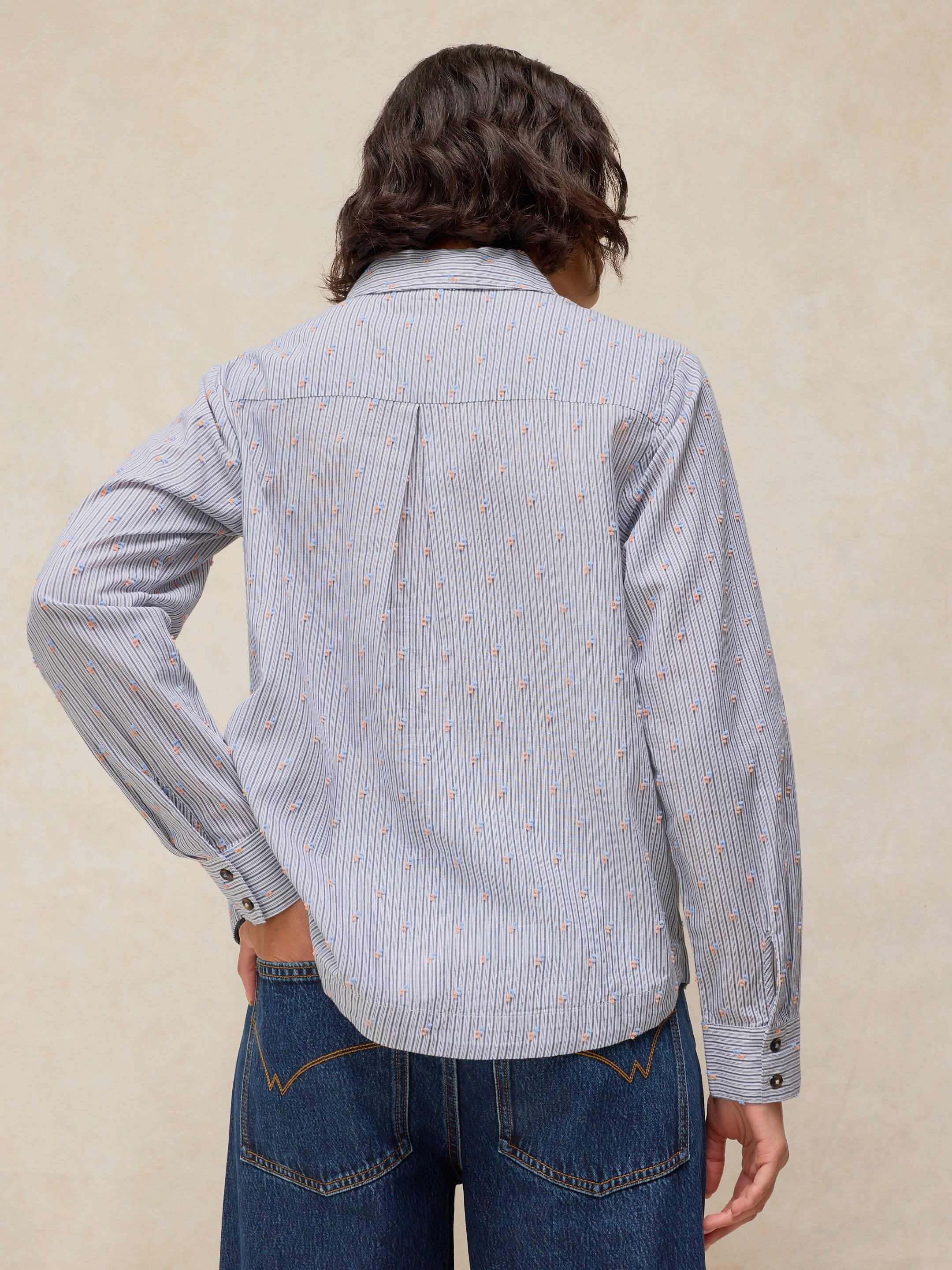 A person shown from the back wearing a light blue and white vertically striped button-up shirt with a small orange pattern, paired with blue jeans, against a textured beige background.
