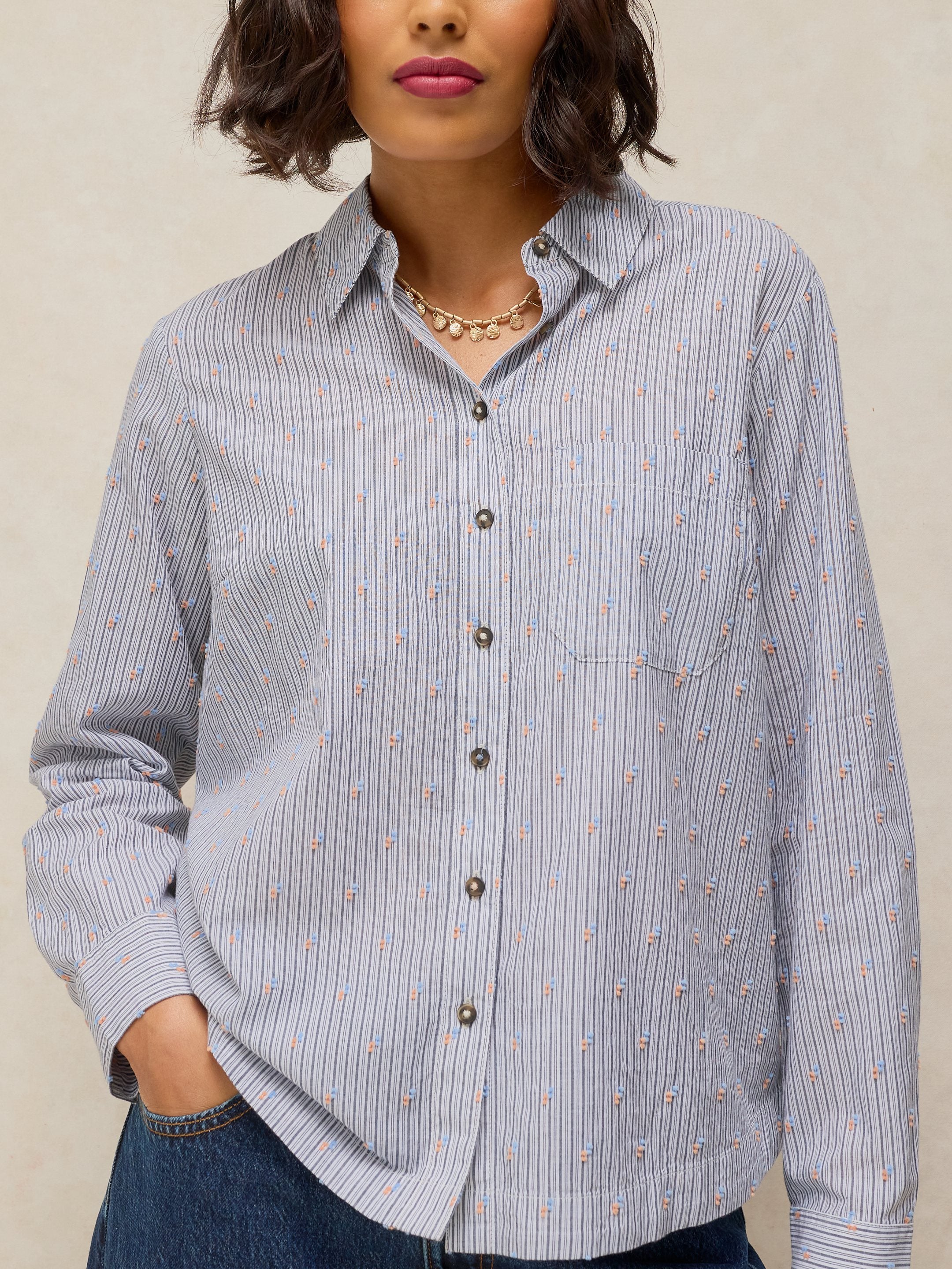 A woman wearing a light blue and white vertically striped button-down shirt with small pink floral details, paired with dark blue jeans and a gold necklace.