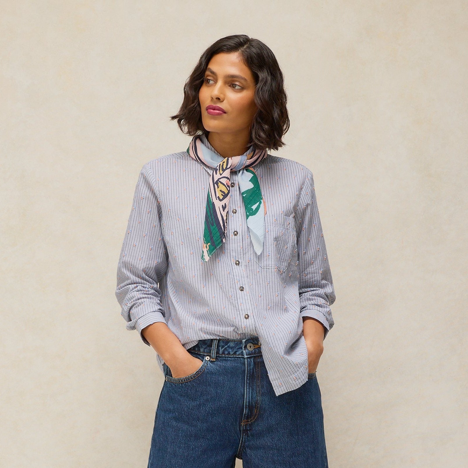 A woman modeling a light blue pinstriped button-down shirt, wide-leg blue jeans with rolled cuffs, a green and white patterned neck scarf, and brown leather loafers, set against a neutral beige textured studio backdrop.
