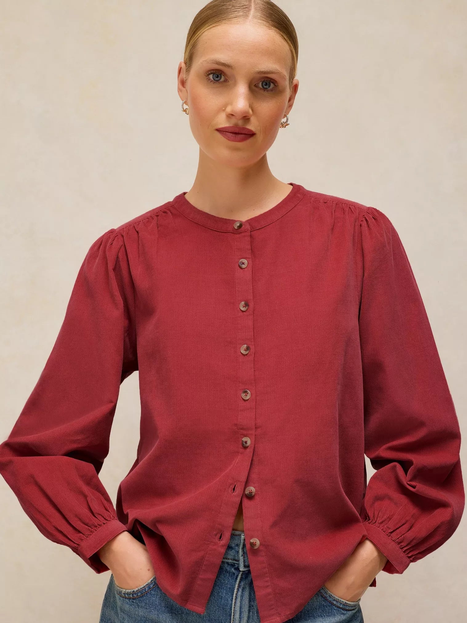 A woman wearing a long-sleeve button-front blouse in a deep burgundy red, paired with blue jeans, against a neutral beige background.