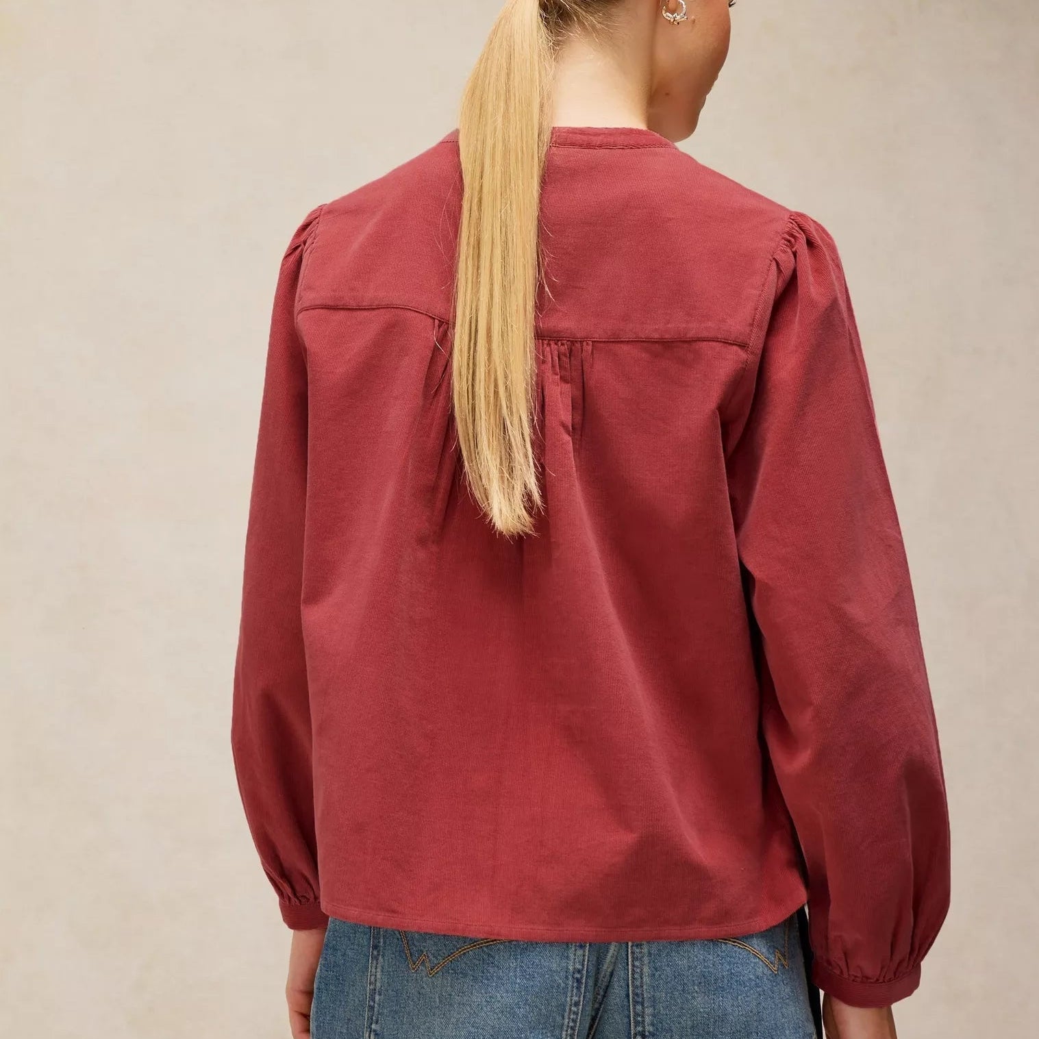 A woman viewed from the back wearing a dusty red long-sleeve blouse with gathered shoulders and blue jeans, standing against a neutral beige textured wall.
