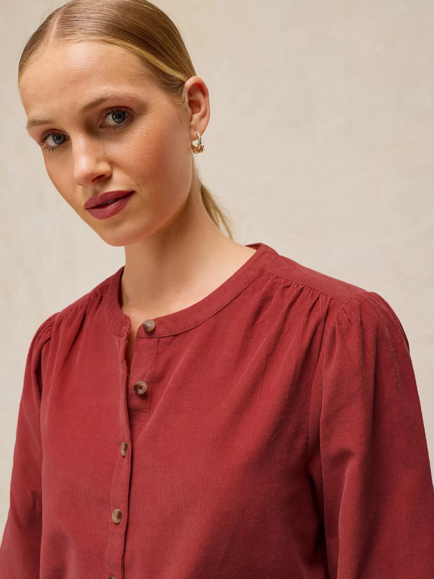 A close-up shot of a woman wearing a rust-red long-sleeve blouse with a buttoned round neckline, set against a neutral beige background.