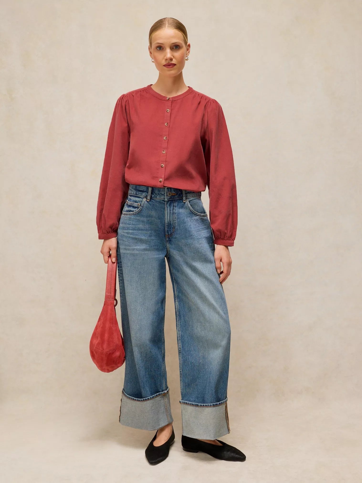 A woman stands against a textured beige studio backdrop wearing a muted red long-sleeve blouse with front buttons, wide-leg blue jeans with rolled cuffs, black flats, and holding a red handbag.