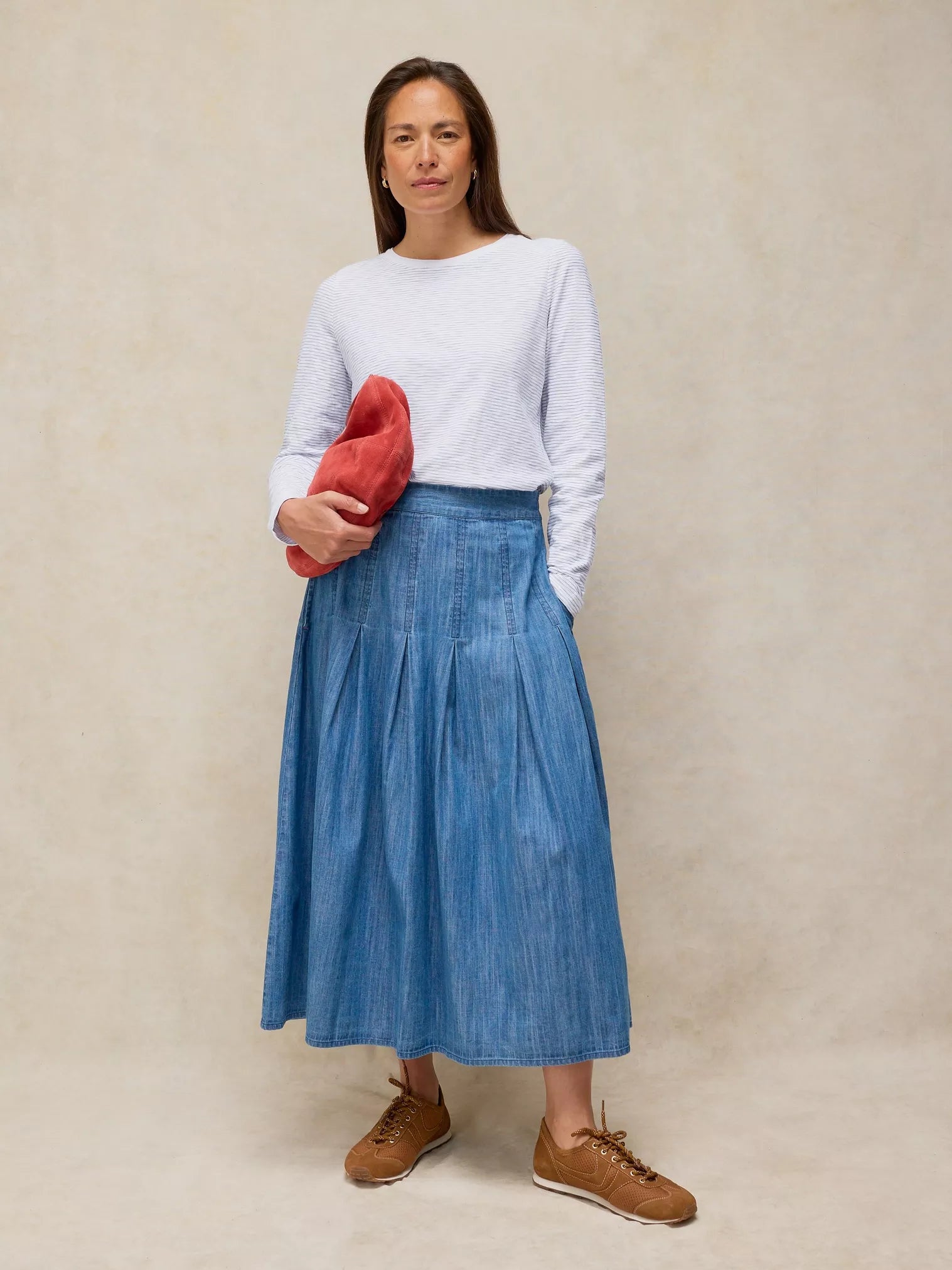 Woman wearing a white sweater and blue denim skirt holding a red clutch against a beige background