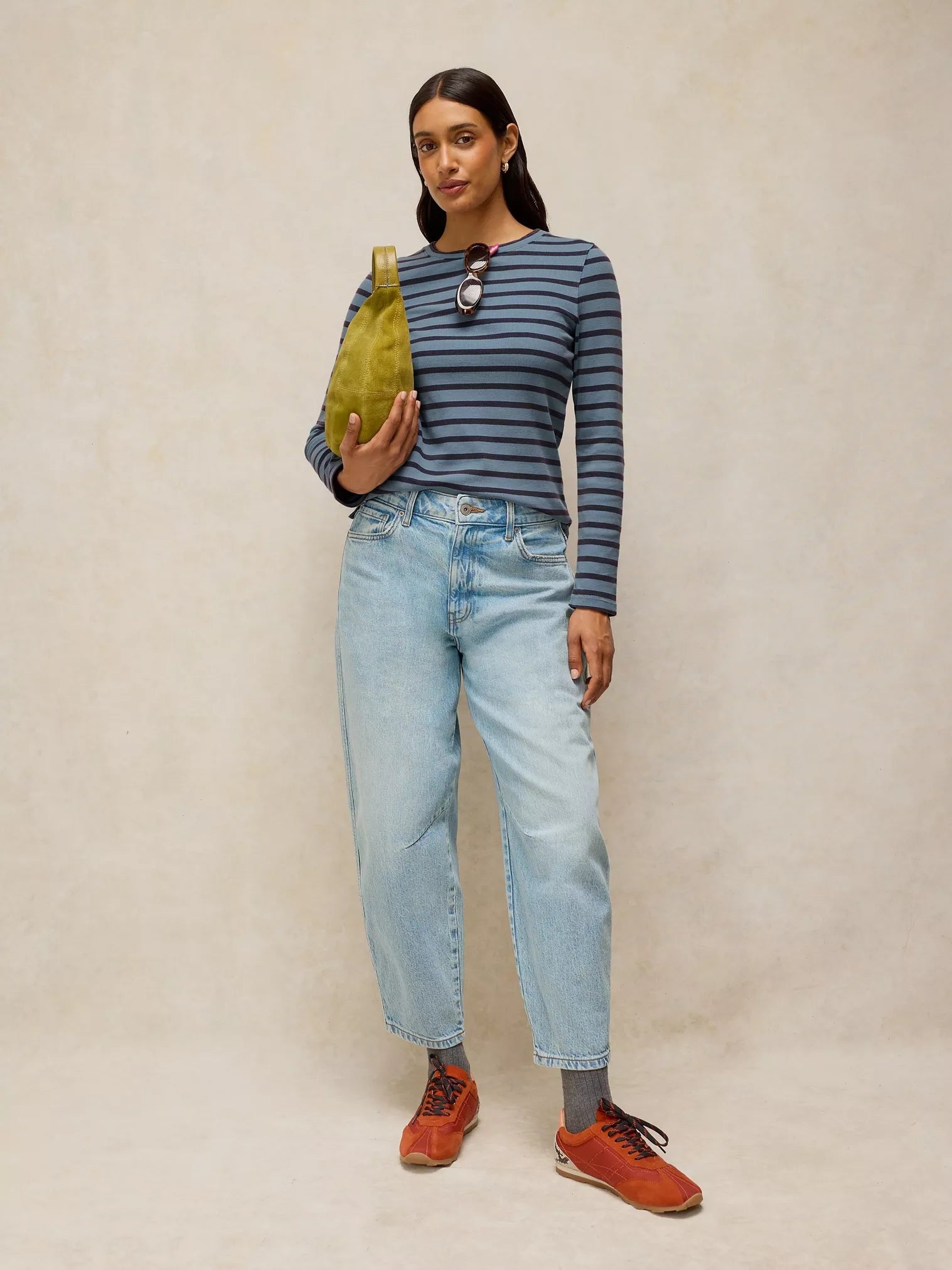 A woman stands against a warm beige studio backdrop wearing a navy and gray striped long-sleeve top, light-wash blue jeans, rust-orange sneakers, gray socks, and holding a small olive green bag.