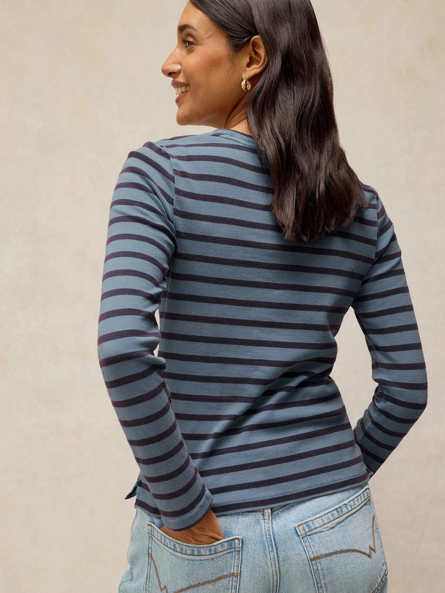 A woman wearing a slate blue and dark gray striped long-sleeve top, shown from a back-three-quarter view against a neutral beige background.