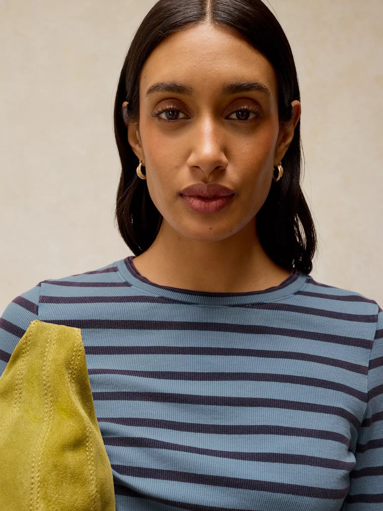 A woman with dark hair wearing a light blue and dark navy striped top, holding a yellow-green textured bag against a neutral beige background.