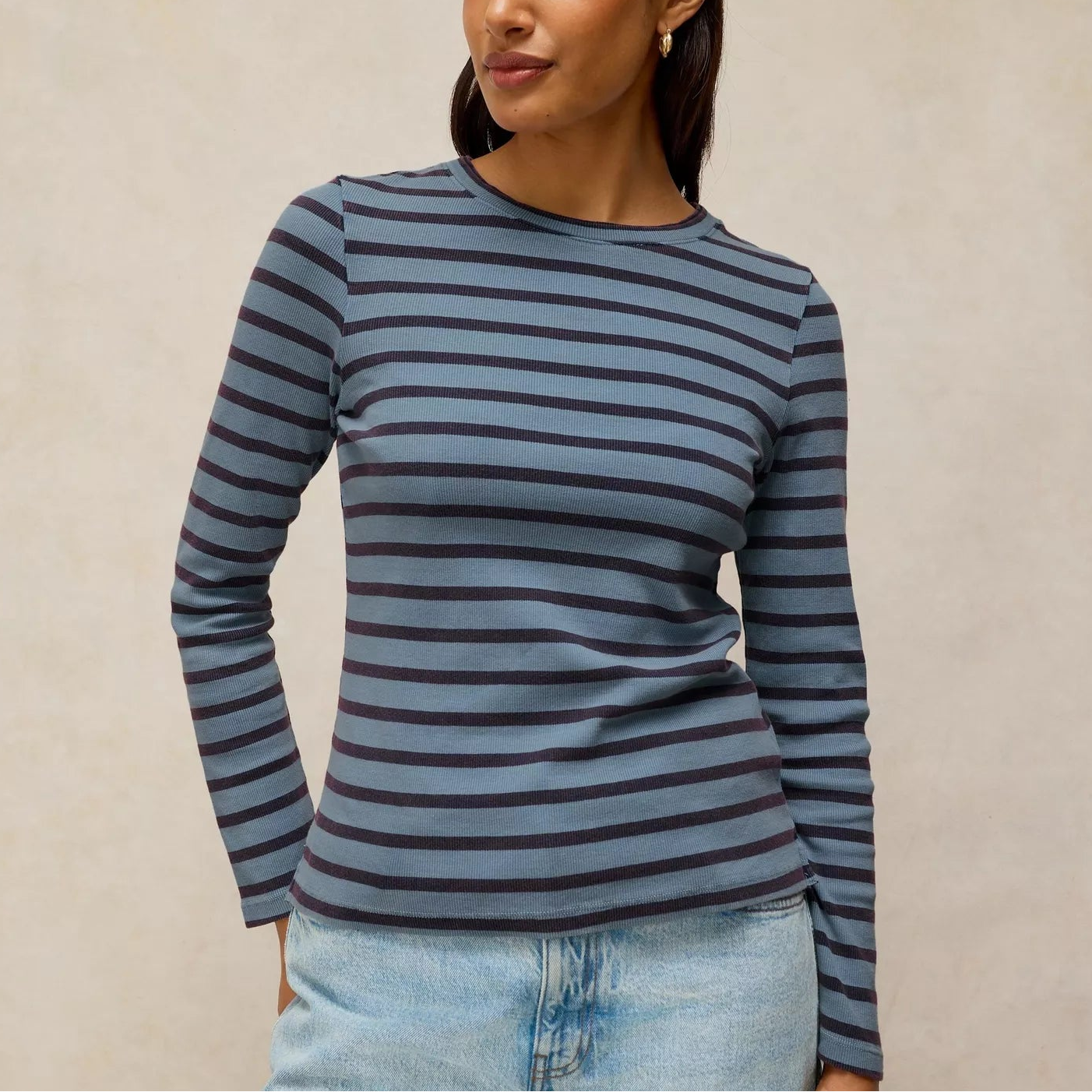 A woman wearing a long-sleeve crewneck top with horizontal navy and gray stripes, paired with light blue jeans, standing against a textured beige background.