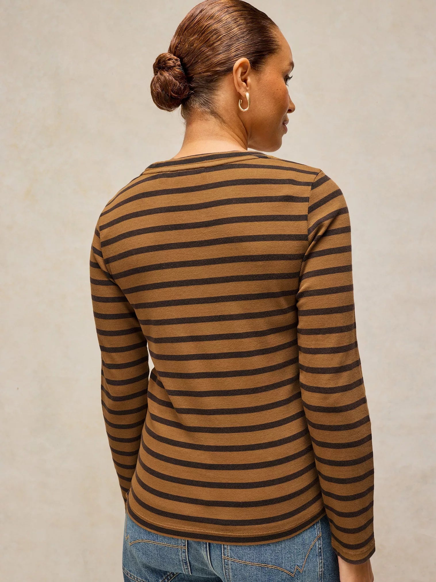 A woman shown from the back wearing a brown and black horizontally striped long-sleeve top, paired with blue jeans, against a neutral beige background.