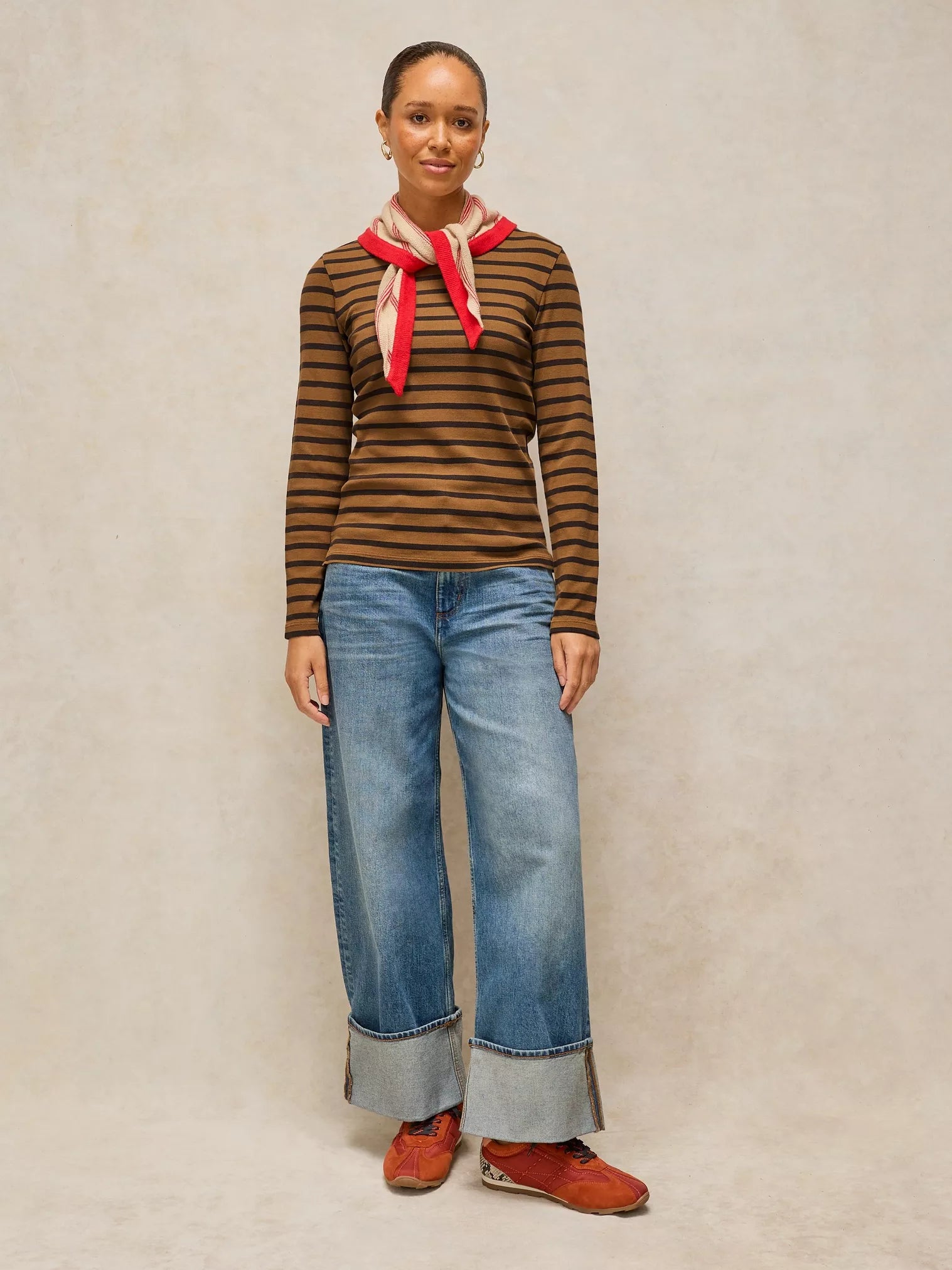 A model stands against a textured beige studio backdrop wearing a brown and black striped long-sleeve top with a red neck scarf, wide-leg blue jeans with rolled cuffs, and brown suede shoes.