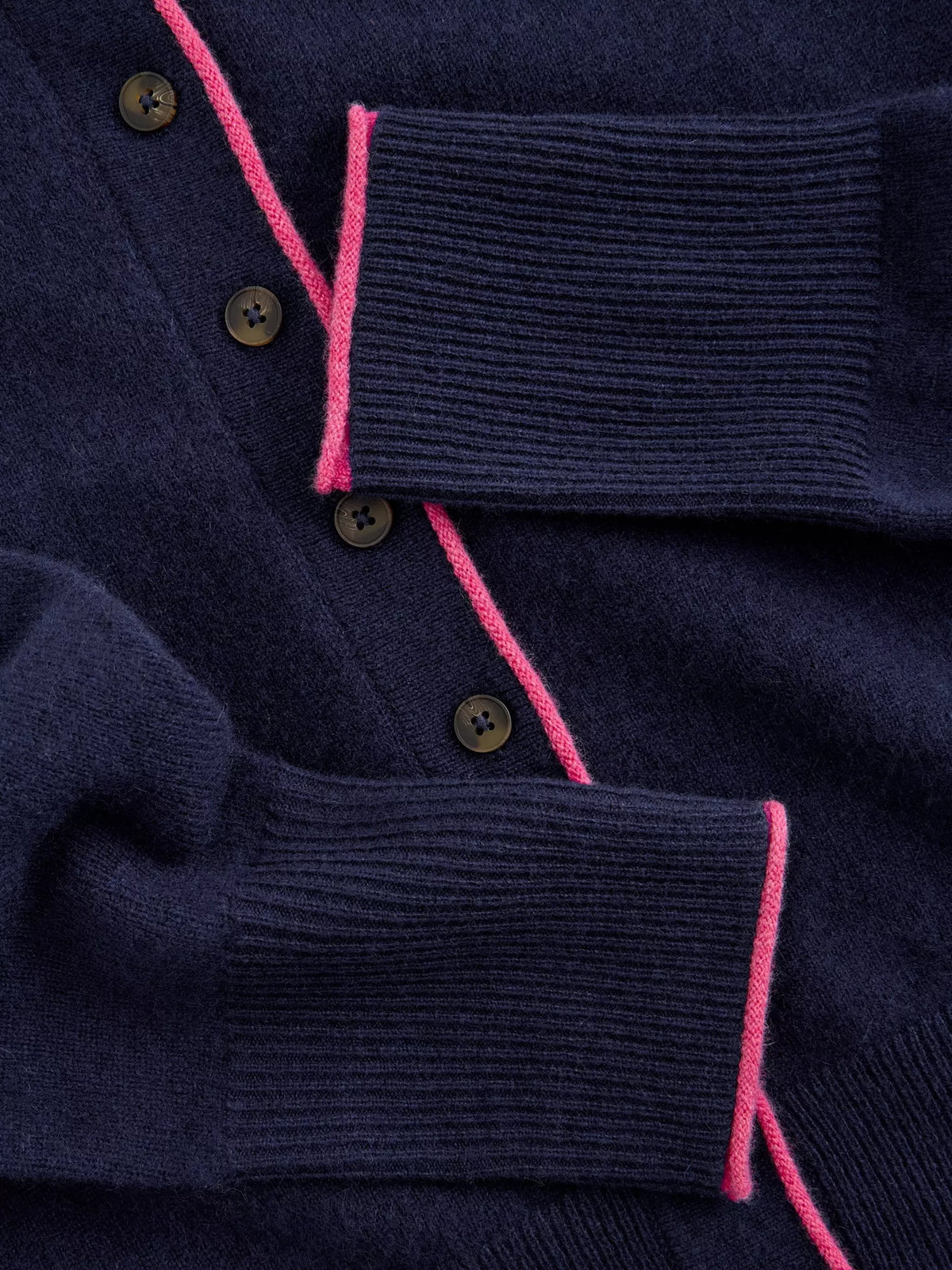 Close-up of a navy blue knit cardigan sweater showing the ribbed cuffs and collar, dark brown buttons, and bright pink contrast stitching.