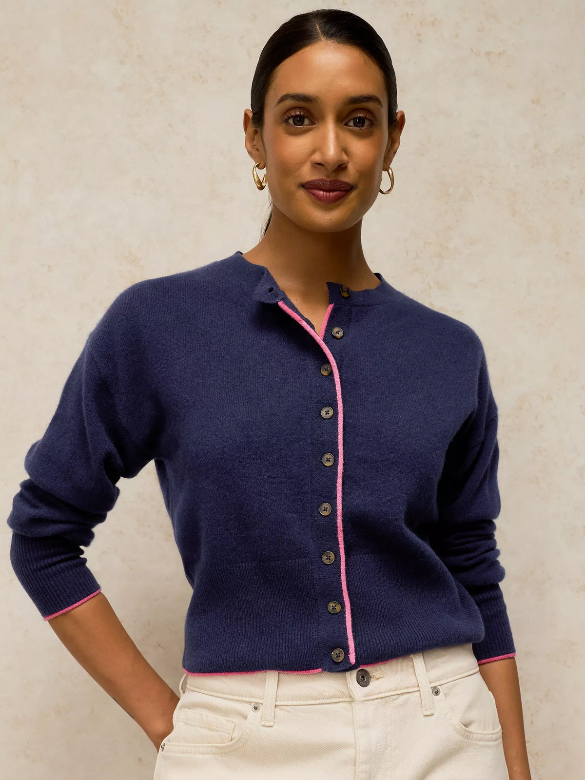 A woman wearing a navy blue knit cardigan with pink trim and buttons, paired with off-white pants, standing against a textured beige wall.